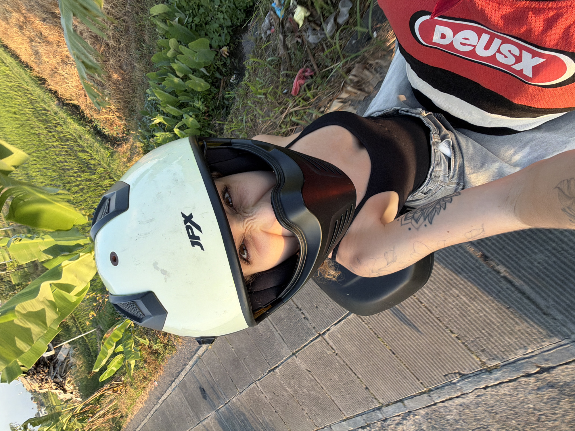 An individual is taking a selfie from a seated position on a scooter in North Kuta, Indonesia. The person is wearing a light green and black helmet, a black tank top, and light blue denim shorts. Their right arm is extended towards the camera, and a portion of their left leg, adorned with tattoos, is visible. Part of a red and black patterned garment, possibly shorts or pants, is also visible. The background features a pathway of grey stone slabs on the left, and on the right, a field with green and brown vegetation, likely rice paddies, interspersed with larger green leaves from banana trees. Some debris and trash are scattered along the right edge of the frame. The lighting suggests it is daytime, likely late afternoon or early morning, given the shadows.