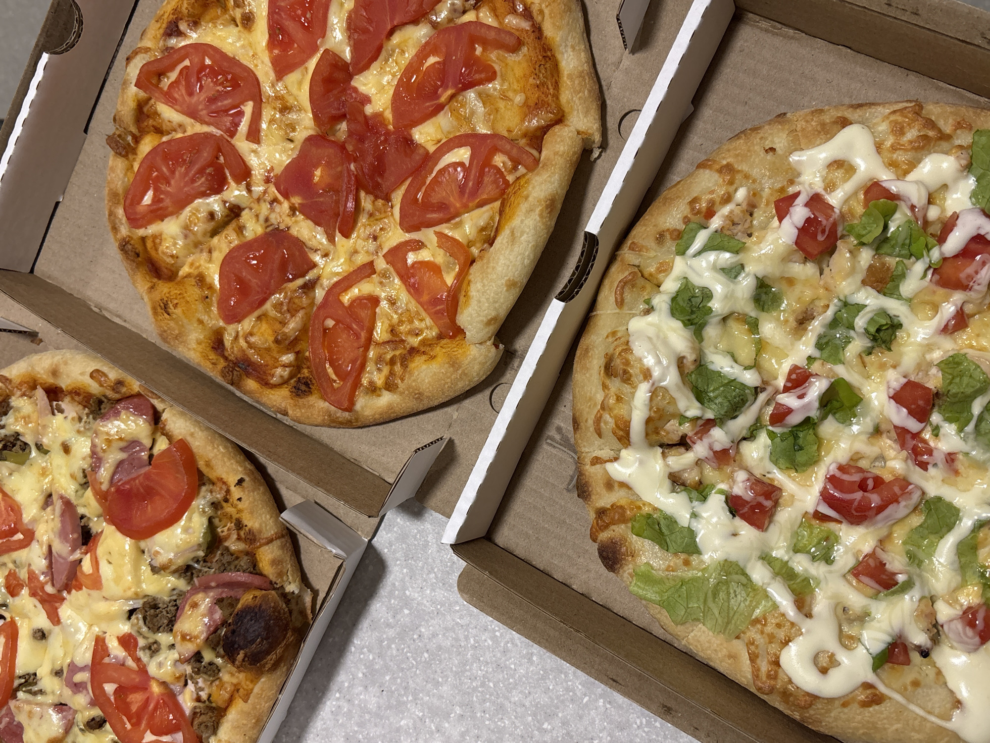 The image shows three pizzas in cardboard boxes on a table. One pizza is topped with tomato slices and cheese. Another pizza has tomato slices, green leaves, and white sauce on top of cheese. The third pizza contains tomato slices, cheese, ham, and what appears to be ground meat. The image was taken in Perm, Russia.
