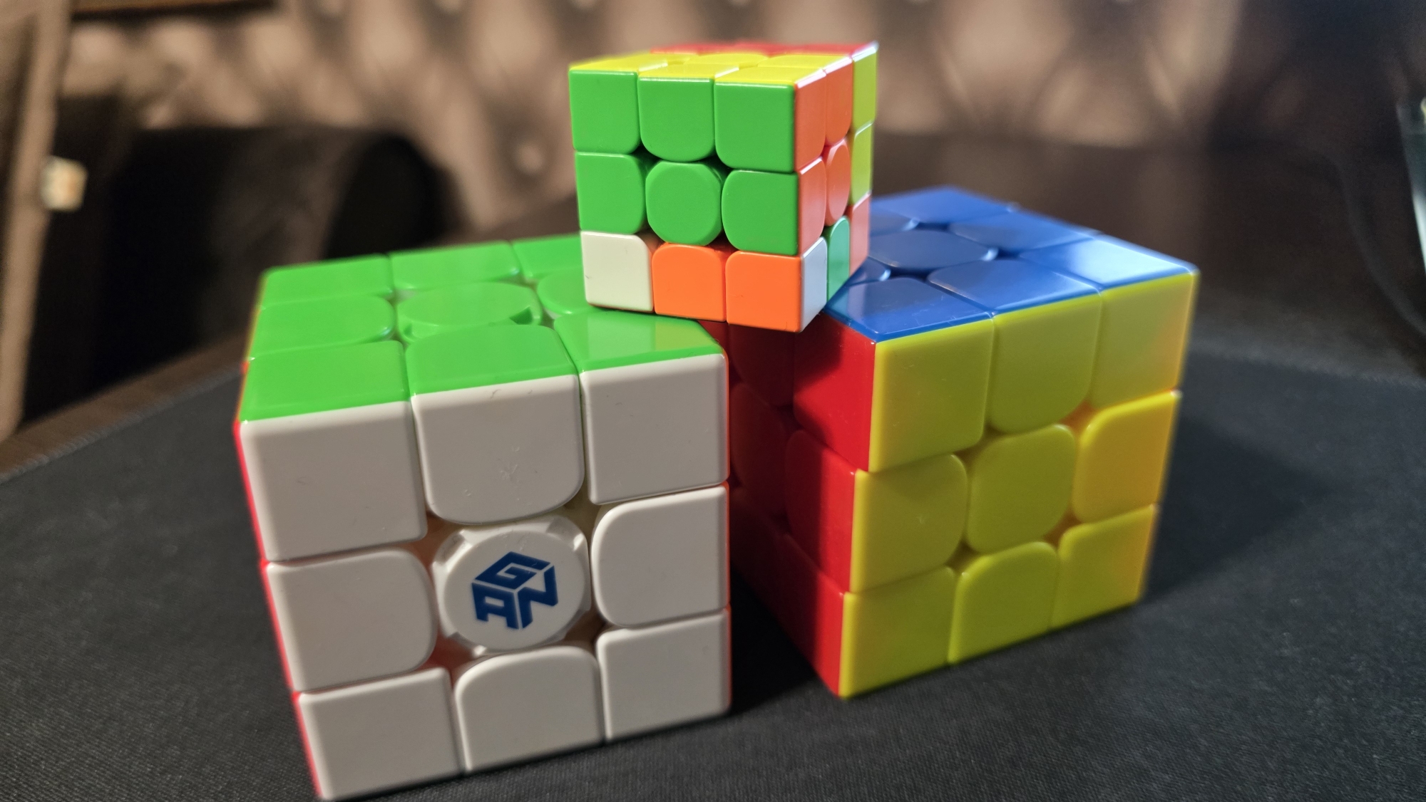 Three Rubik's Cubes are arranged on a black surface, possibly a desk mat. The cube in the foreground left is predominantly white with green and red sides visible. It features a blue logo with the letters "GAN". To the right and slightly behind is another cube, primarily yellow with red and blue sides. A smaller cube sits on top of the yellow and red cube, showing green, orange, and yellow faces. The background is blurred, revealing a dark, textured surface that appears to be upholstery, potentially from a chair or sofa. The lighting casts shadows on the black surface. There are no people or animals present. The scene is an indoor, still-life arrangement of puzzle toys.