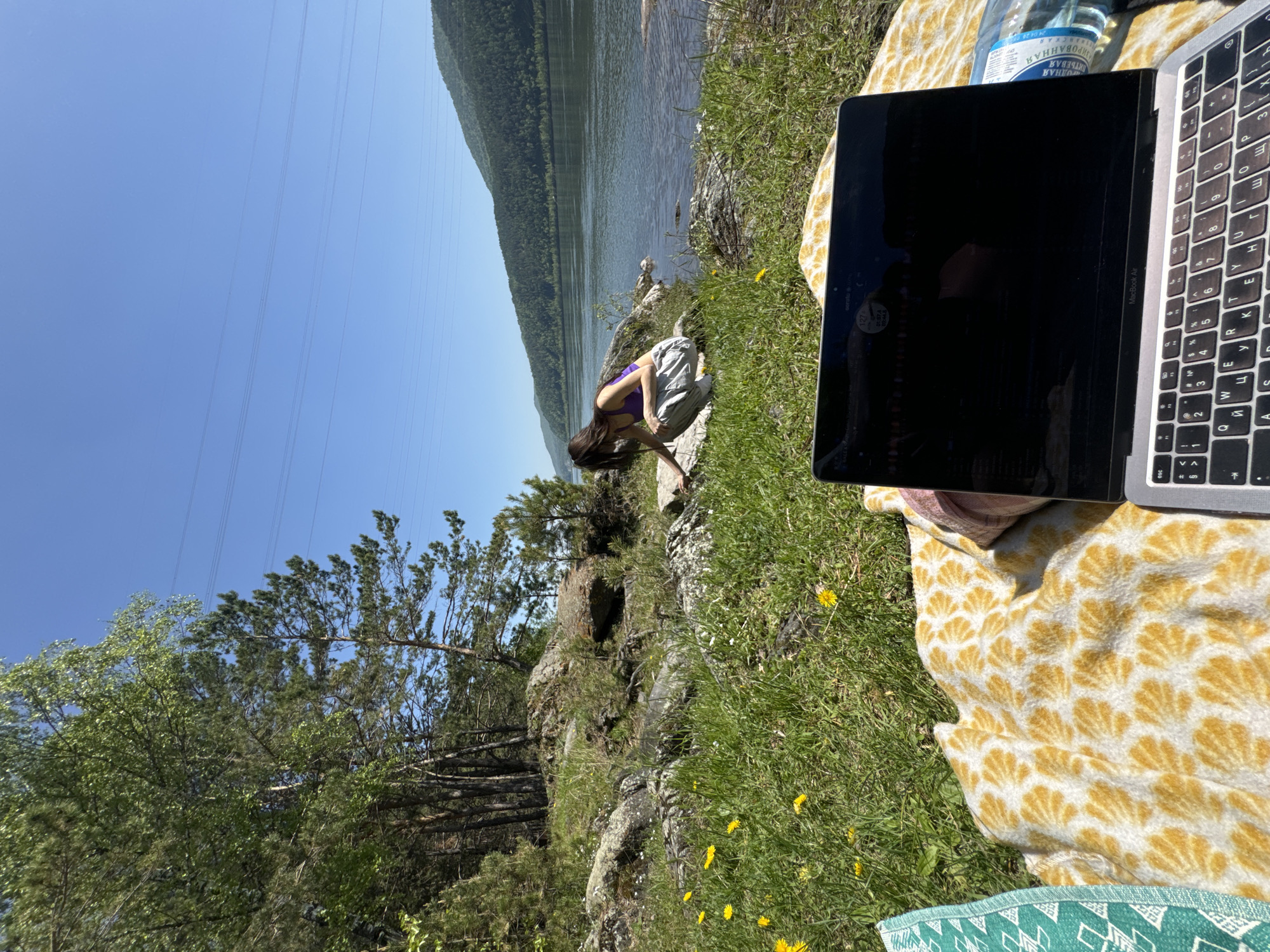 This image captures an outdoor scene by a large body of water, likely a river, on a bright, sunny day. The clear blue sky is visible, with several power lines stretching across it. The landscape includes a sloping, rocky bank covered in green grass and small yellow flowers (dandelions), leading down to the water. Across the expansive body of water, densely forested mountains or large hills rise in the distance.
In the foreground, a yellow and white patterned blanket is spread on the grass, partially covering a green and white geometric-patterned cushion or blanket at the bottom. A silver MacBook Air laptop with a Cyrillic keyboard rests open on the yellow blanket, its screen dark. Adjacent to the laptop is an inverted plastic bottle of water, labeled with Cyrillic text that reads "РОДНАЯ ПИТЬЕВАЯ ГАЗИРОВАННАЯ" (Rodnaya Drinking Carbonated), along with a date stamp "24.04 20 0".
Further back, near the water's edge, a young woman with long dark hair, wearing a purple top and light-colored pants, is crouching on the rocky ground. She appears focused on something close to the water or the bank. Various trees, including conifers and deciduous types, dot the landscape, particularly on the left side of the image.
The scene conveys a sense of tranquility and a connection with nature, suggesting a leisurely outdoor activity or a remote workspace. The bright sunlight indicates it is daytime, possibly morning or early afternoon. The Cyrillic text on the laptop and water bottle, combined with the natural scenery, aligns with the location context of Слизнево, Russia.