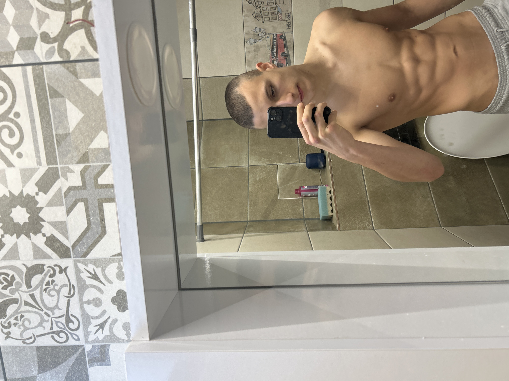 The image shows a shirtless young man in gray shorts taking a selfie in a bathroom. He is holding a black smartphone in his right hand, and his face is visible in the reflection. The bathroom has beige tile flooring and walls, with decorative tiles featuring building and bus designs. A toilet is visible in the background. A blue bottle, toothpaste, and a cleaning sponge are on the floor. The selfie is taken in front of a framed mirror surrounded by decorative tiles. It is not possible to determine the exact location in Burgas, Bulgaria, without further information.