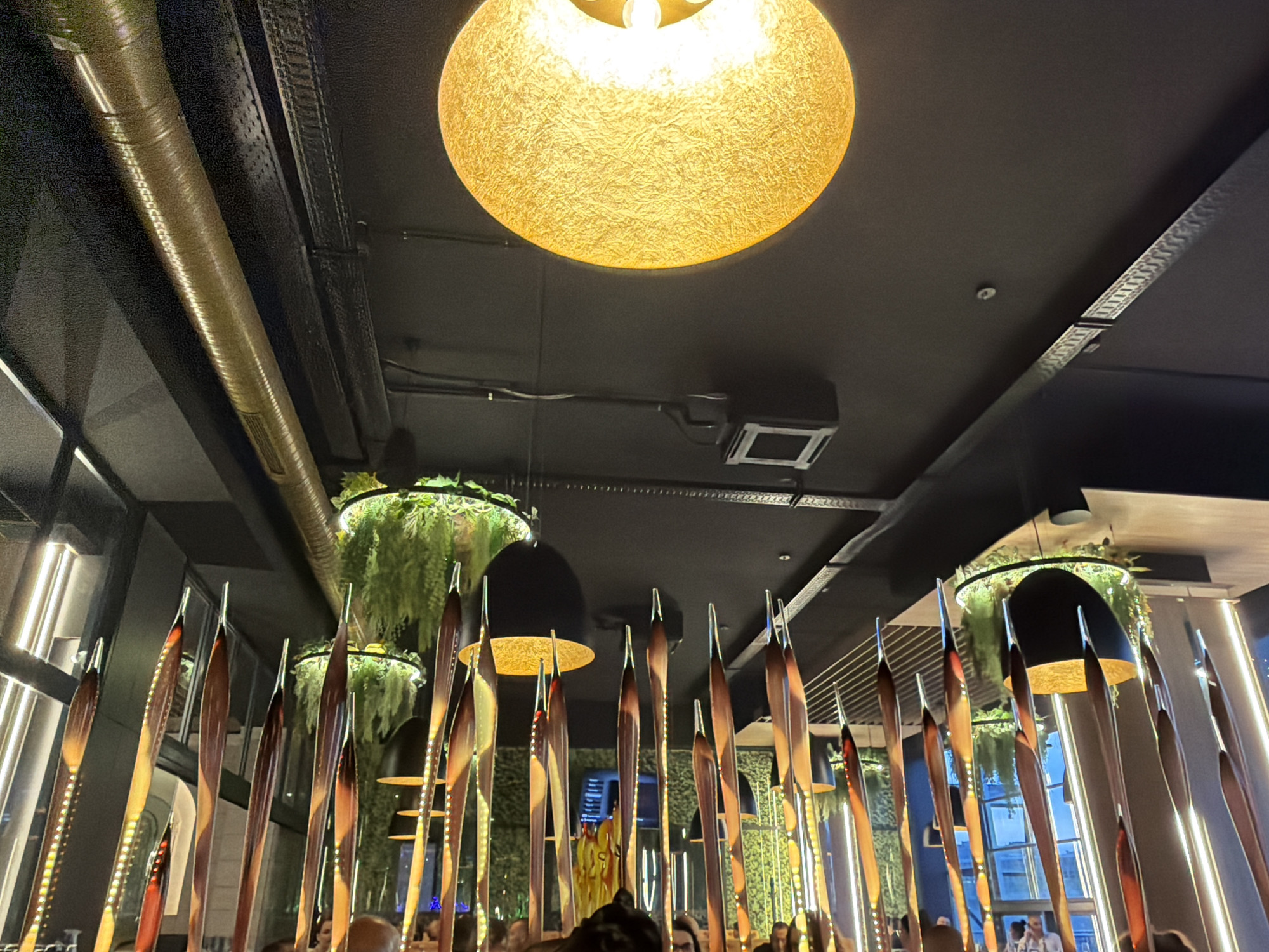 This image captures an indoor scene, likely a restaurant, bar, or lounge, characterized by its modern and decorative ambiance. The perspective is looking upwards towards a dark, possibly black, ceiling from beneath the main decorative elements.
The ceiling features exposed industrial-style components, including a prominent golden, corrugated pipe running diagonally, black conduits, and track lighting. Several light fixtures are visible. In the foreground, a large, textured, spherical or irregularly-shaped pendant light glows warmly with a golden-yellow hue. Further back, several smaller, dark, bowl-shaped pendant lights are suspended, some revealing golden interiors. A square ventilation or light fixture is also integrated into the ceiling, alongside a small, round sensor or smoke detector.
A striking feature of the decor is a series of tall, narrow, elongated decorative elements, resembling stylized leaves or blades, which are arranged in rows in the midground. These elements have a polished, reflective surface, showing shades of brown, gold, and red, and appear to have internal or adjacent lighting creating a dotted pattern along their length.
Green, lush hanging plants are suspended in circular planters from the ceiling, adding a natural and vibrant touch to the space. The background reveals large windows or reflective surfaces, showing blurred linear strip lighting and hints of an outside environment or reflections of the interior.
In the lower portion of the image, numerous people are visible, mostly obscured by the decorative elements or blurred due to motion or depth of field. They appear to be gathered, socializing, or dining, consistent with a lively evening or nighttime event. The overall lighting scheme and the presence of people suggest a social gathering in a sophisticated setting. No discernible text is visible in the image.