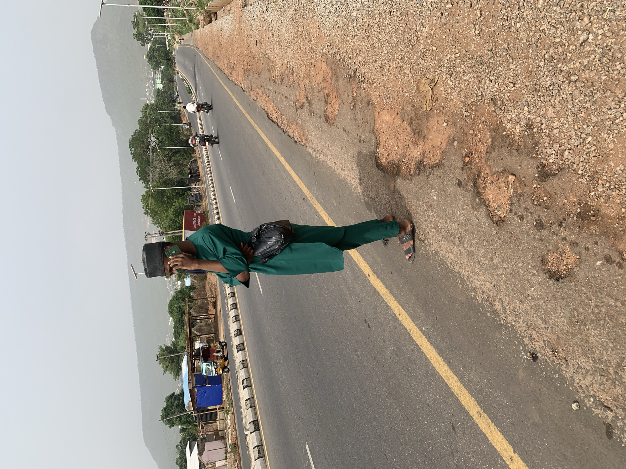 A medium shot presents a person on the shoulder of a road in Adankolo, Nigeria, identifiable by the setting. The person, wearing green attire and a black cap, is looking at their phone. A black bag is slung across their body. The road is gray asphalt with a yellow centerline stripe. On the road, motorcycles are visible, moving away from the viewer. The right edge of the frame reveals a shoulder of loose gravel and dirt. On the left side of the road, structures, possibly buildings, are present. In the distant background, a mountain is visible. The sky is overcast.
