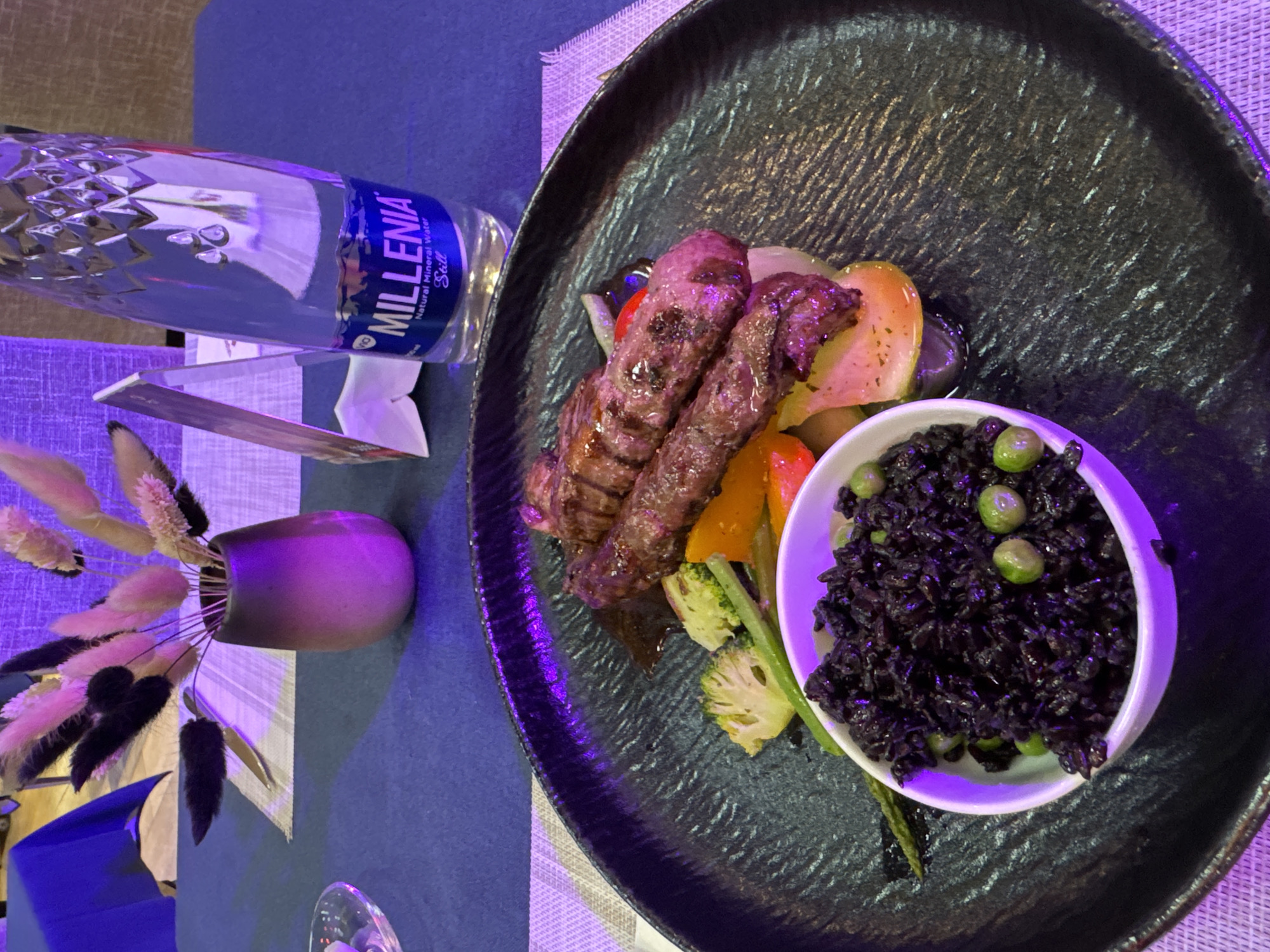 The image shows a plate of food consisting of grilled meat, vegetables, and black rice with peas, placed on a textured black plate. A bottle of Millenia water is visible next to the plate, along with a small vase containing dried flowers. The setting appears to be indoors, likely a restaurant, with a blue tablecloth and a patterned placemat. While the image suggests a dining scene, there is no specific information indicating it is in Ulaanbaatar, Mongolia. Additional visual or contextual information is required to confirm the location.