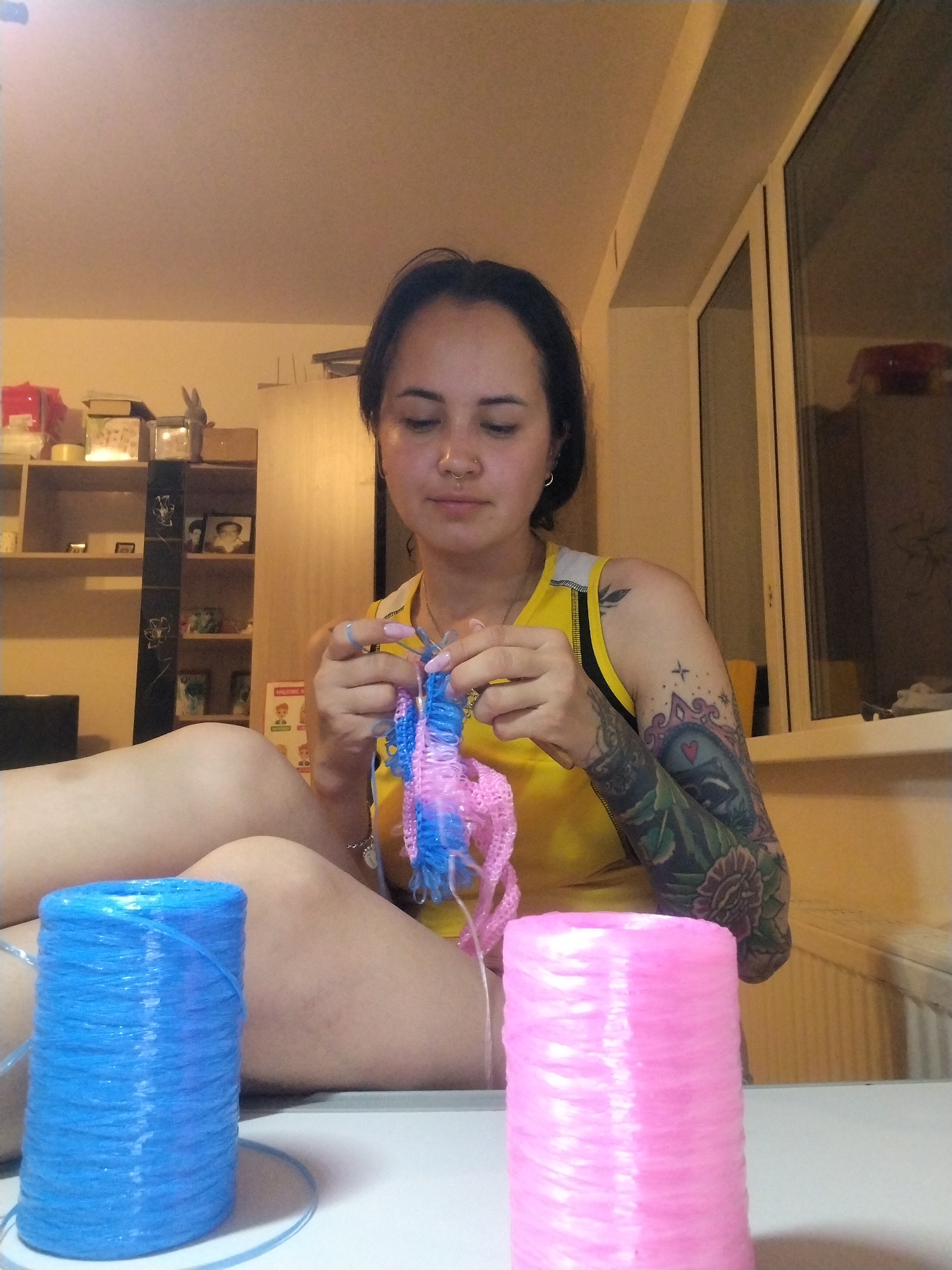 A young woman is knitting with blue and pink plastic twine in an indoor setting. She has dark hair pulled back, a nose ring, and is wearing a yellow tank top. A large tattoo covers her right arm. Two spools of twine, one blue and one pink, are on a white surface in front of her. The background includes a shelf with various objects, a window, and a light-colored wall. The location (city of Noviy, Russia) cannot be determined from the image.
