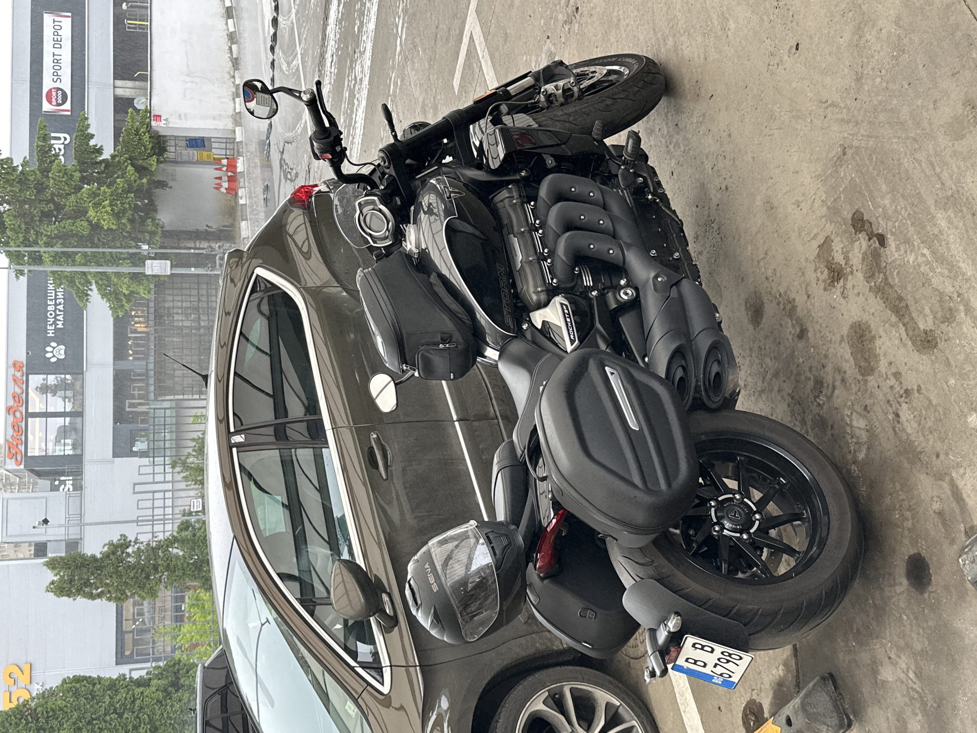 The image depicts a motorcycle parked next to a car in a parking area in Veliko Tarnovo, Bulgaria. The motorcycle is a black Triumph Rocket 3 Storm with luggage bags attached, parked at an angle relative to the parking space lines. A car, likely a dark-colored Opel, is parked adjacent to the motorcycle on its right. The motorcycle has a license plate reading "BB 86798." A black motorcycle helmet is resting on the rear of the bike. The environment is an outdoor parking area with surrounding commercial buildings, including a "SPORT DEPOT" storefront and a building with Cyrillic signage, "HE4OBELIKII MAGA3UH." The ground is concrete with stains and tire marks.