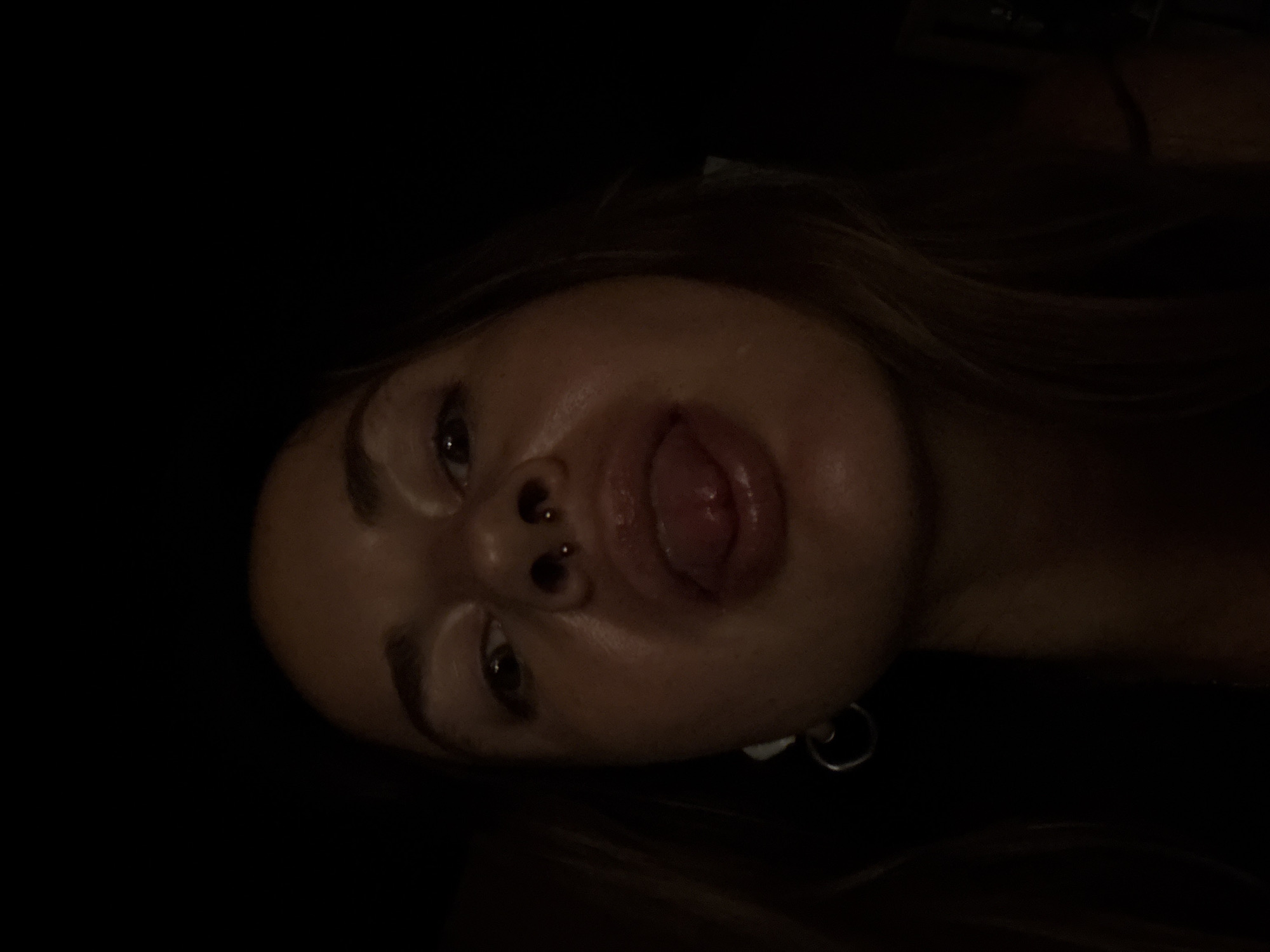 A low-angle, close-up selfie of a young woman is taken in a dimly lit environment. The woman is looking directly at the camera with her tongue extended. She has a septum piercing and is wearing a hoop earring on her left ear. Her skin appears moist, and there is a slight sheen on her face. The background is almost entirely dark, with a hint of her hair visible on the right side. The image is grainy due to the low light conditions.