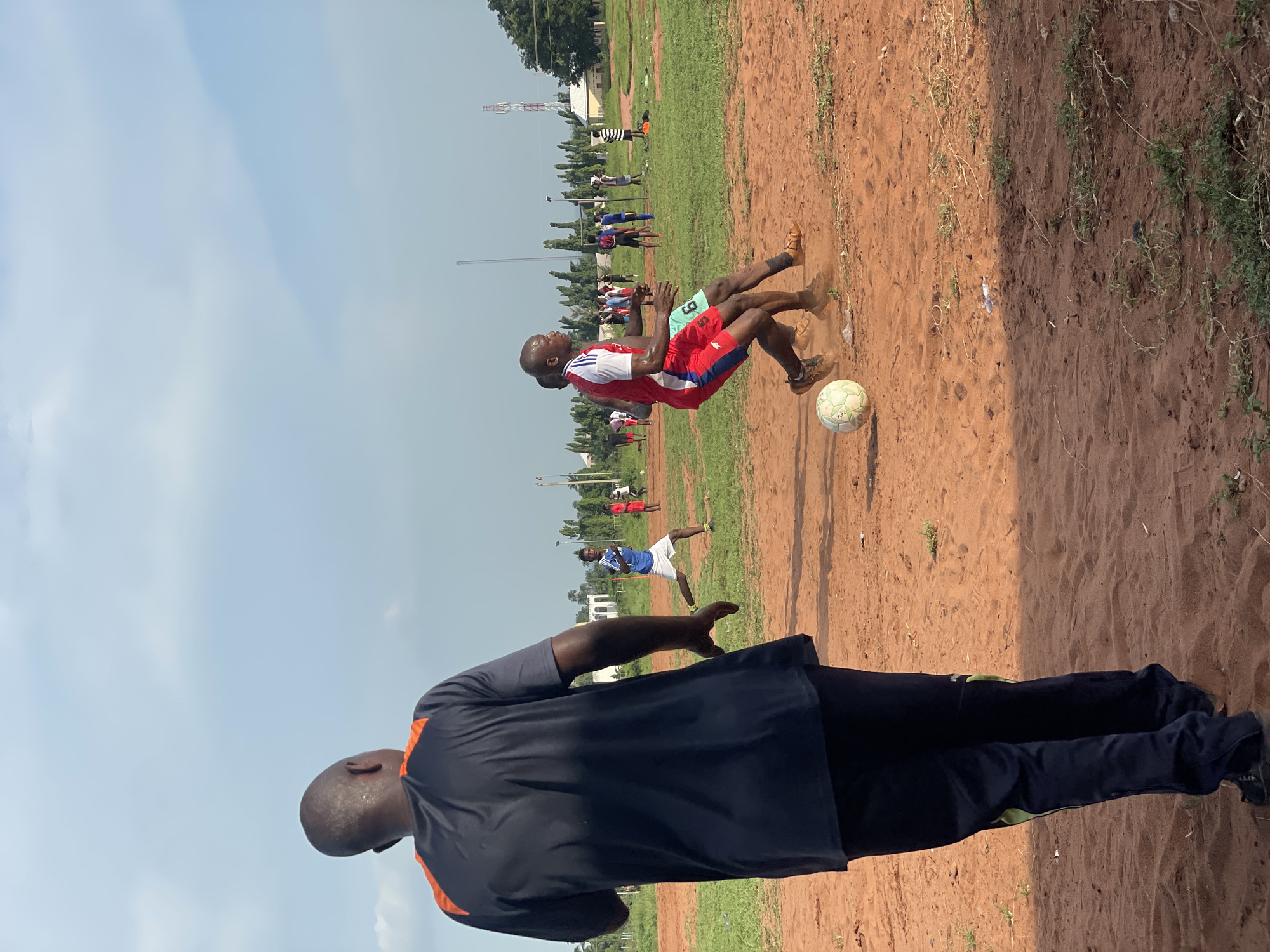 The image captures an energetic football match or practice taking place on a sunlit, unpaved field in Ogbogbo, Nigeria. In the foreground, a person, seen from behind, wears a dark blue or black t-shirt with orange shoulders and dark pants, observing the play intently.
The central action unfolds in the mid-ground, where two players are actively engaged. One player, wearing a red and white striped jersey with the number "6" visible on his back and blue shorts, is caught mid-air in an acrobatic pose, possibly attempting an overhead kick or a high volley. A cloud of reddish dust emanates from the ground around his feet, indicating dynamic movement on the sandy pitch. Another player, in a light blue or green shirt and red shorts, is positioned nearby, also in motion. A white football is visible close to the feet of the player in the red and white jersey.
The playing surface is predominantly reddish-brown dirt with scattered patches of green grass. In the background, several other players are visible, some standing, some engaged in the game, suggesting a full team or group participation. Beyond the field, a line of tall, slender trees provides a natural border, while buildings and a prominent cell tower stand against a clear, light blue sky. The bright daylight suggests it is midday or early afternoon, with strong shadows cast across the field. The scene depicts an informal yet passionate sporting event under an open sky.