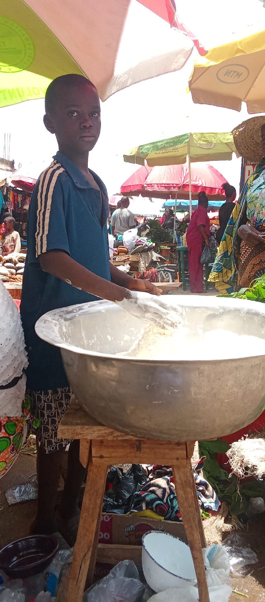 In a bustling outdoor market in Akwanga, Nigeria, a young boy stands behind a large, silver-colored metal bowl perched on a wooden stool. He wears a blue t-shirt with white stripes on the shoulders and patterned shorts. His hands are inside the bowl, appearing to be mixing a white substance. The market scene behind him includes people shopping under brightly colored parasols, piles of goods, and various vendors. Discarded items and trash are visible near the base of the stool, including plastic bags and bowls.