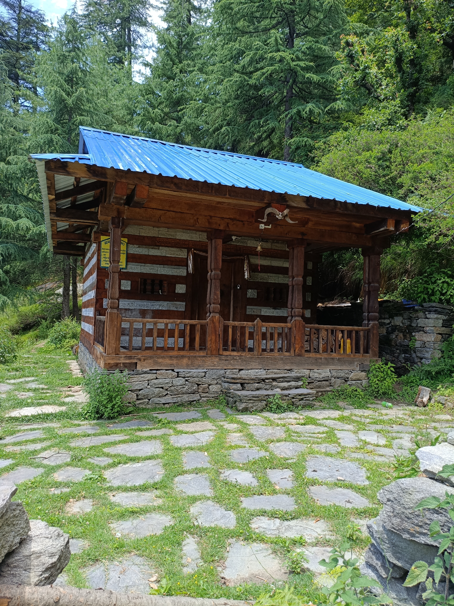 The image depicts a small, wooden building with a blue metal roof, nestled within a dense forest. The building has a covered porch with wooden pillars and a railing. The walls appear to be constructed of horizontal wooden logs alternating with stone. A stone pathway, with grass growing between the stones, leads up to the entrance of the building. The surrounding environment is lush and green, with tall trees visible behind the structure. There are no people present in the image, and the scene appears to be tranquil and natural.