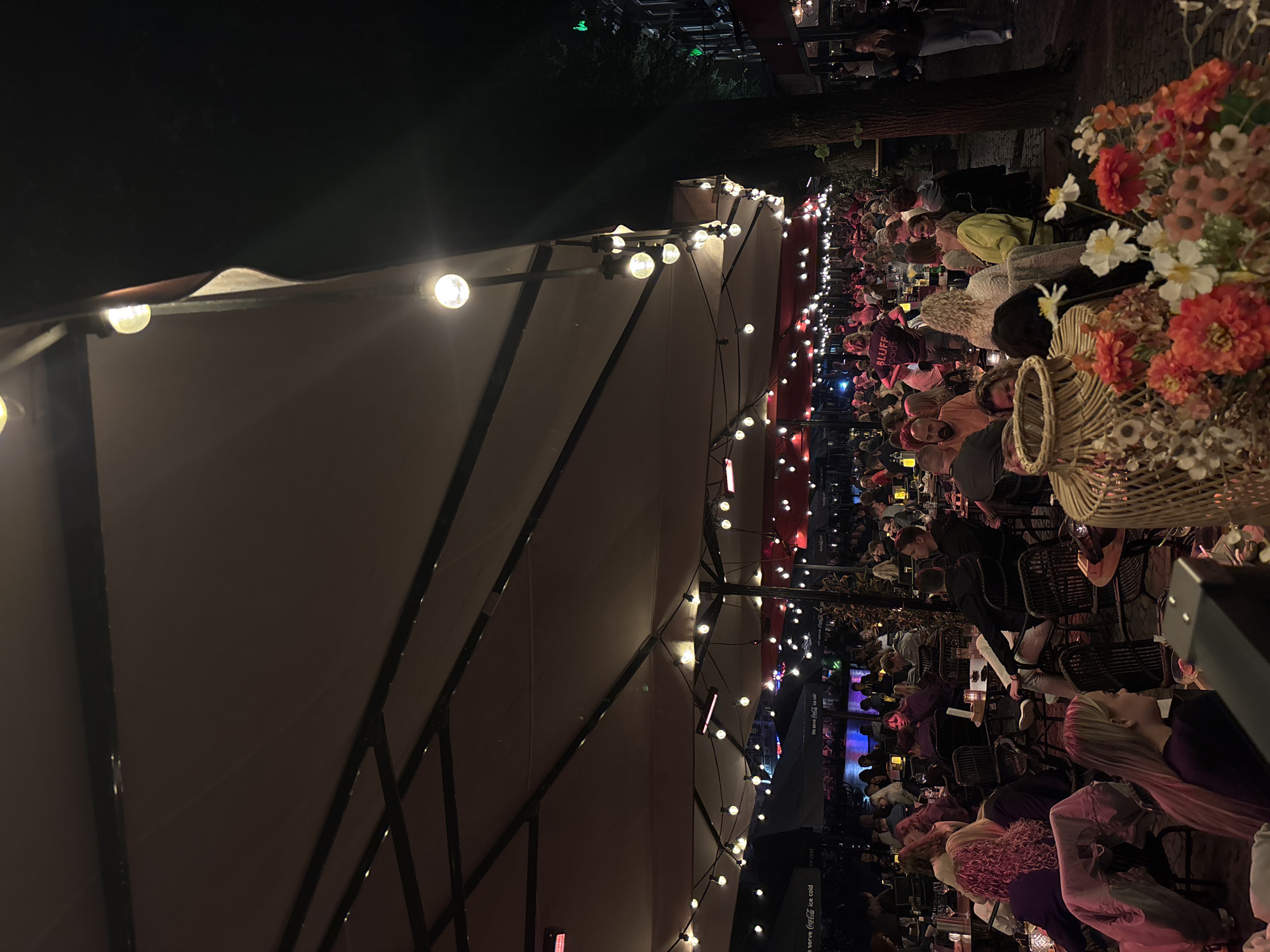 The image depicts an outdoor dining area at night, likely a restaurant or cafe, illuminated by string lights and overhead heaters. People are seated at tables, engaged in conversation and dining. The scene suggests a lively atmosphere with a moderate crowd. In the background, there appears to be a body of water, possibly a canal or river, with reflections of lights. The architecture visible suggests an urban setting. The location is identified as being in Enschede, Netherlands.