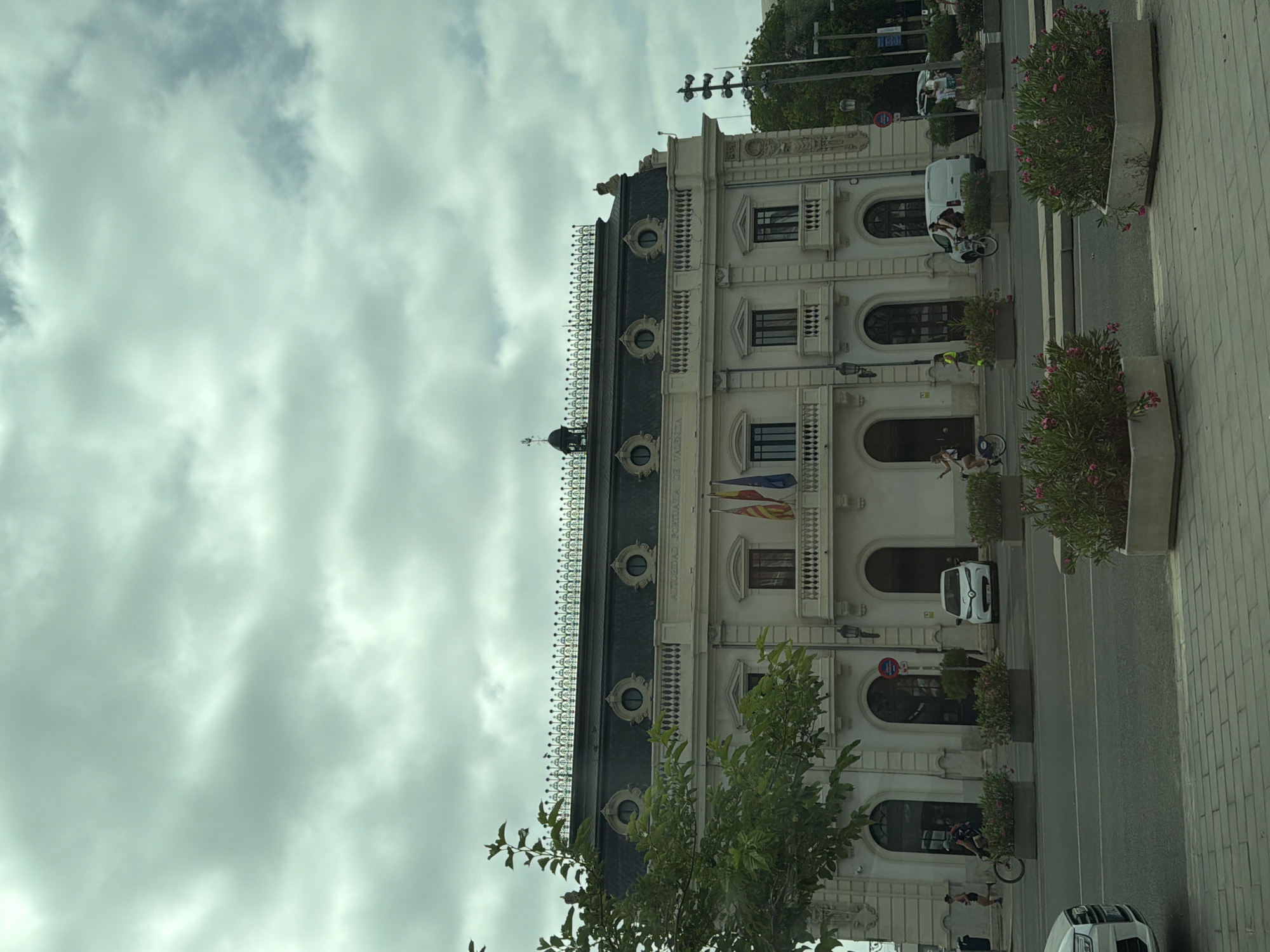 The image shows a street scene in front of an ornate, light-colored building with a dark roof. The building has multiple windows, arched doorways, and decorative elements along its facade. There are flags flying on the building.
Several people are visible in the scene. One person is riding a bicycle on the sidewalk. Another person appears to be walking or standing near a parked white van. A few other individuals are seen in the background, some near parked vehicles and others on the sidewalk.
Objects in the scene include parked cars, a white van, and planters with pink flowering plants. The street is paved with cobblestones on the right side. The sky is cloudy, suggesting overcast weather. The lighting indicates it is daytime.
The building's facade has text that reads "AUTORIDAD PORTUARIA DE VALENCIA" vertically. This text suggests the location is related to the Port Authority of Valencia. The overall impression is a daytime urban scene on a cloudy day, with everyday activities taking place in front of a historically significant or architecturally interesting building.