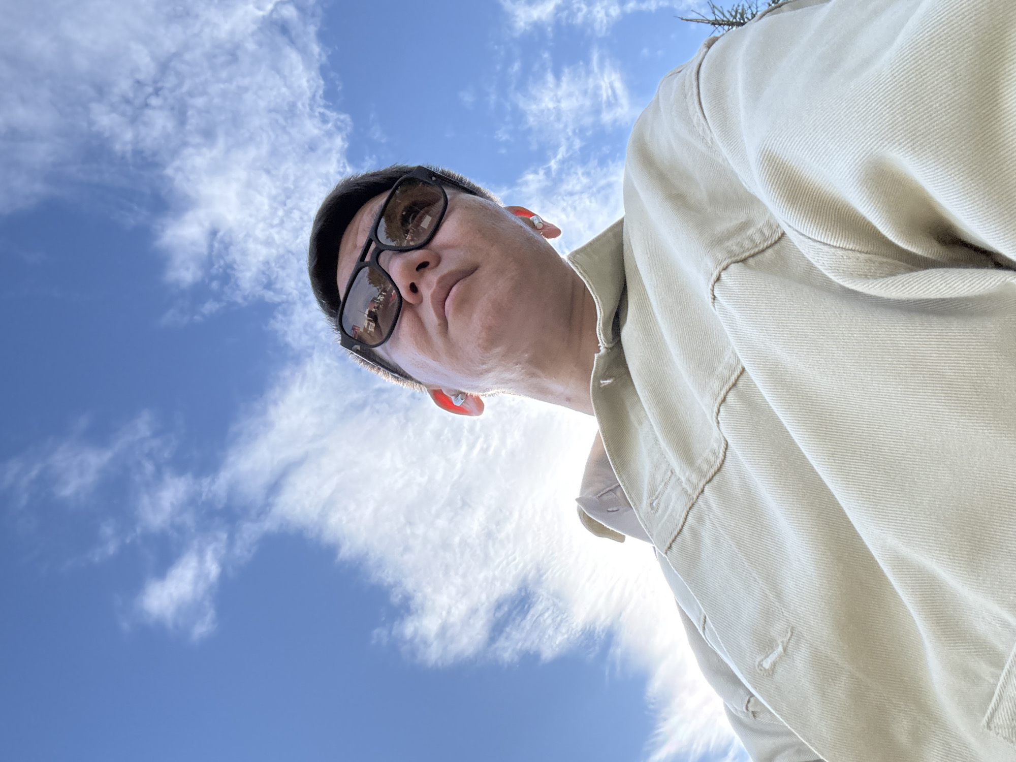 A low-angle shot captures a person wearing sunglasses and a light-colored jacket, looking upwards at a bright blue sky with scattered white clouds. The person has short, dark hair and appears to be wearing earbuds. The jacket is a utilitarian style with visible stitching and buttons. The sky is partly obscured by the person's head and shoulders. No specific location in Moscow, Russia, can be determined from the image.