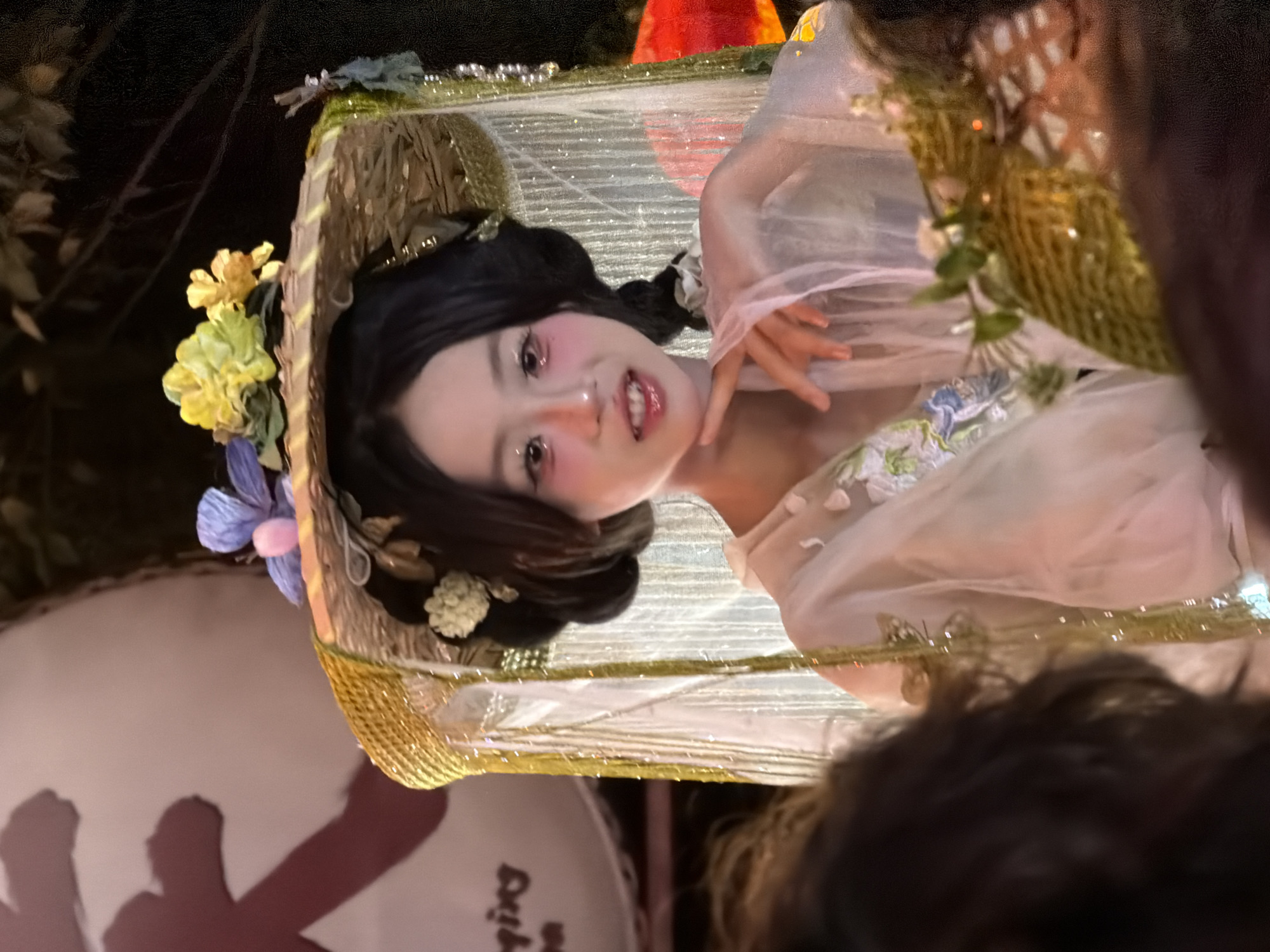 A young woman in traditional Chinese attire, adorned with a floral straw hat and delicate makeup, is the central subject of this photograph, taken at night, likely during a parade or festival in Yuzhong District, China. The woman has dark hair styled in buns, with strands braided. She wears a light, translucent dress with floral embroidery. Her hand is raised, delicately touching her chin, and she has a subtle smile. The straw hat is decorated with blue and yellow artificial flowers, and a sheer, shimmering veil hangs from it. The background is dark, with some indistinct shapes suggesting a festive outdoor setting, possibly illuminated by lanterns. The image captures a moment of elegance and cultural representation.
