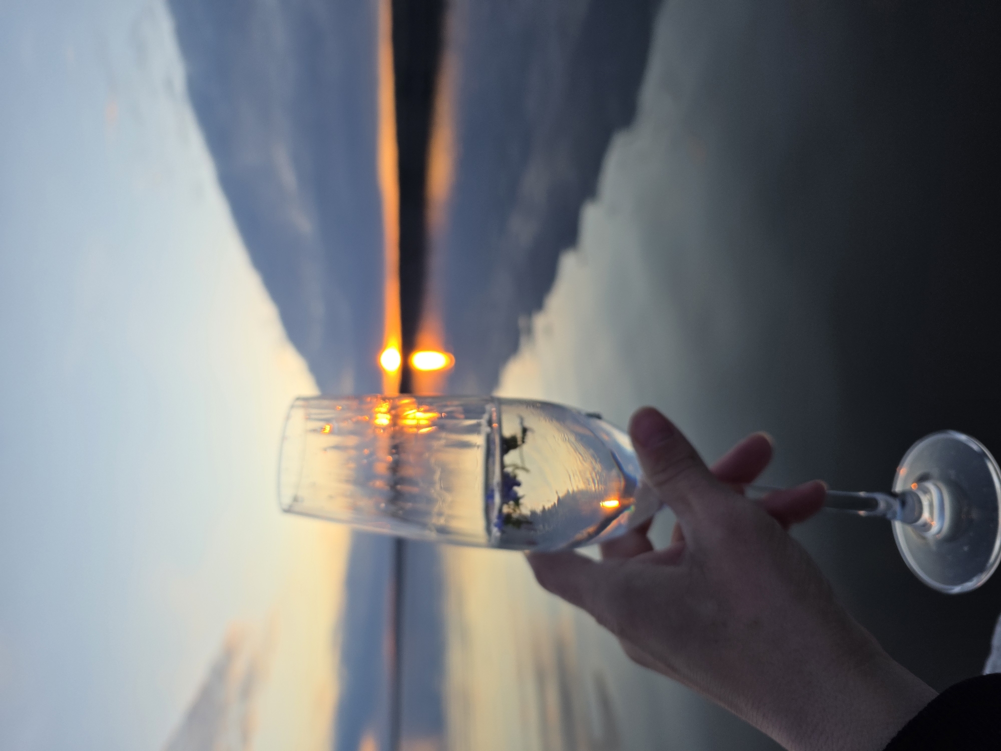A hand holds a champagne glass in front of a sunset over water. The sunset's reflection is visible in the water and in the glass. The photograph was taken in Chaikovsky, Russia.