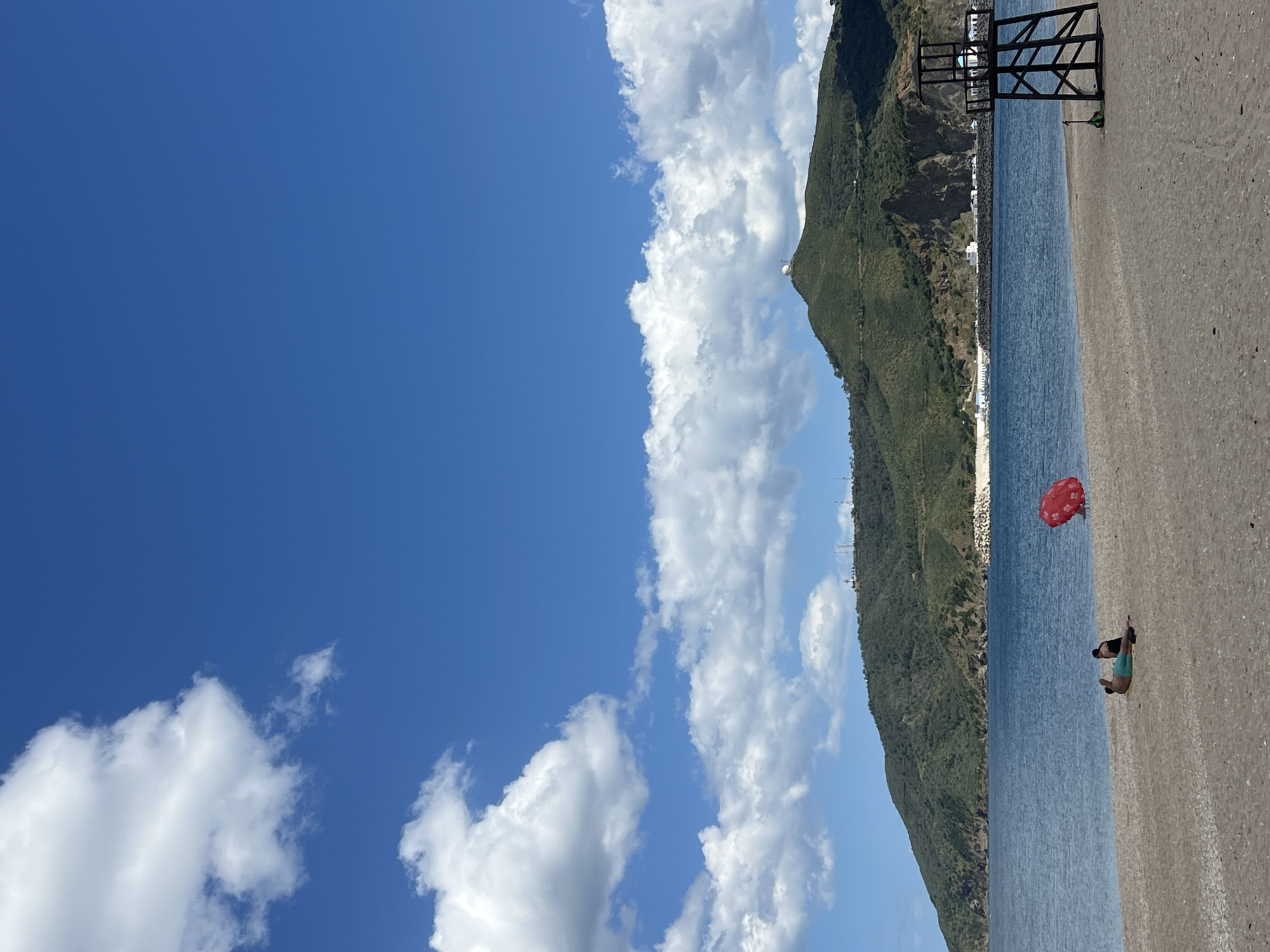 The image shows a beach scene in M'Diq, Morocco. The view is rotated 90 degrees clockwise. The sky is blue with scattered white clouds. A mountain covered with green vegetation runs along the center of the image. The beach is sandy and meets a body of water, likely the Mediterranean Sea.
A person is lying on the beach in the lower right corner, possibly sunbathing. A red umbrella is planted nearby. A black metal structure is near the water’s edge further up the beach. White buildings are visible along the coastline.