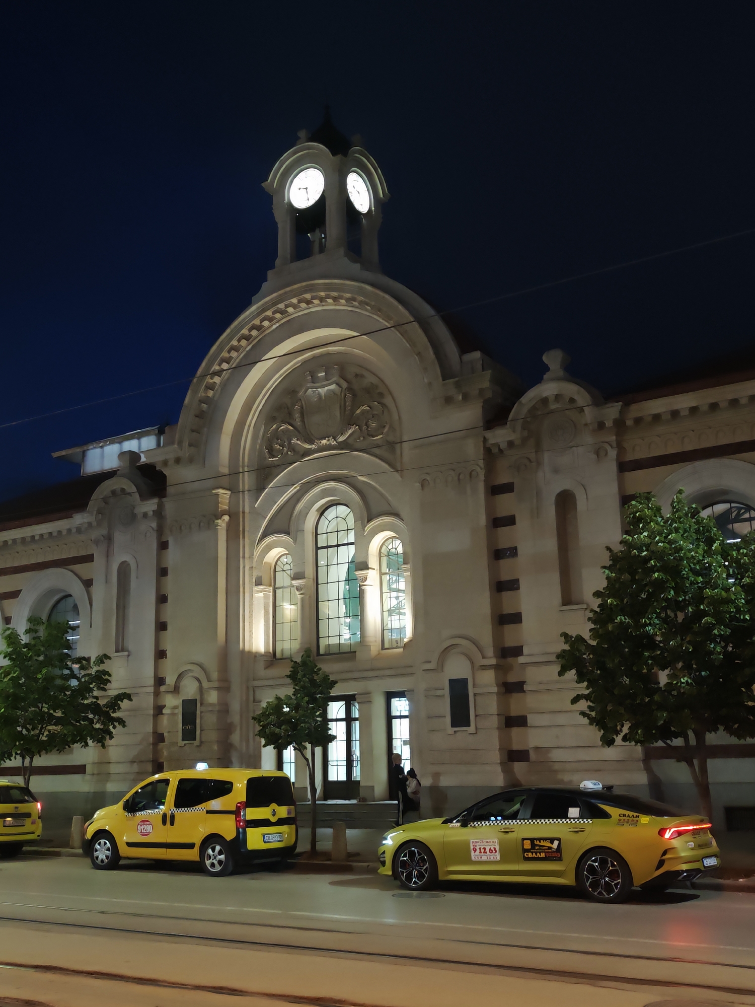 The image shows the Central Market Hall in Sofia, Bulgaria at night. The building is brightly illuminated, highlighting its architectural details including its clock tower, arched windows, and ornate facade. Several yellow taxis are parked in front of the building, with visible markings and numbers. A few people stand near the entrance of the building. The sky is dark blue, suggesting late evening or night. Trees are planted along the sidewalk. The ground indicates a paved road with some tracks or lines embedded in it.