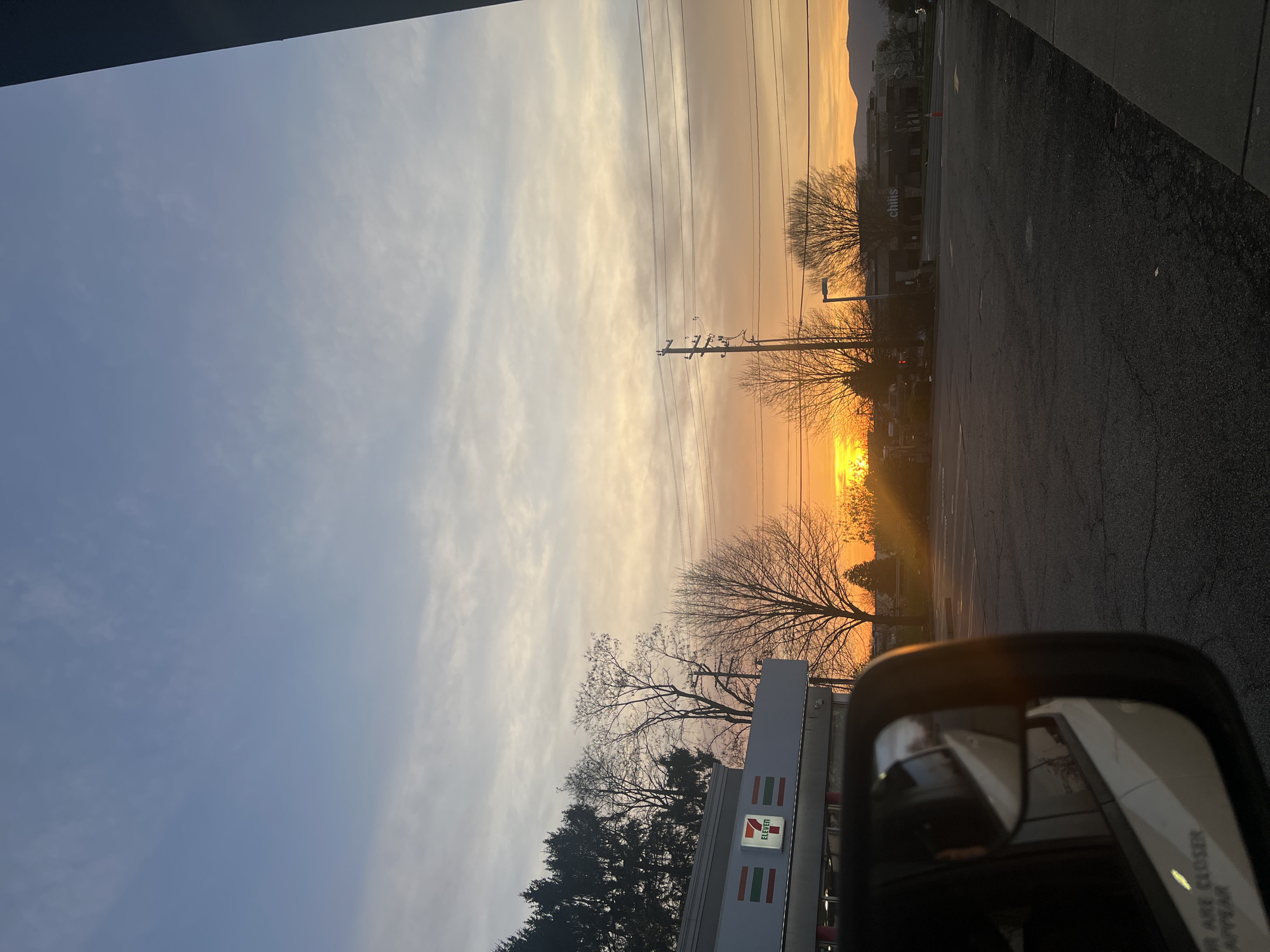 The image captures a sunset scene in what appears to be a commercial area. A 7-Eleven store is visible on the left side, identifiable by its logo. The setting sun casts a bright orange glow on the horizon, silhouetting trees and utility poles. A building that might have a Chili's restaurant is also discernible. A car side mirror is visible in the lower right corner, with the reflection of other vehicles. The photograph seems to be taken from inside a car, and the environment suggests an urban or suburban setting in the City of Newburgh, United States of America.