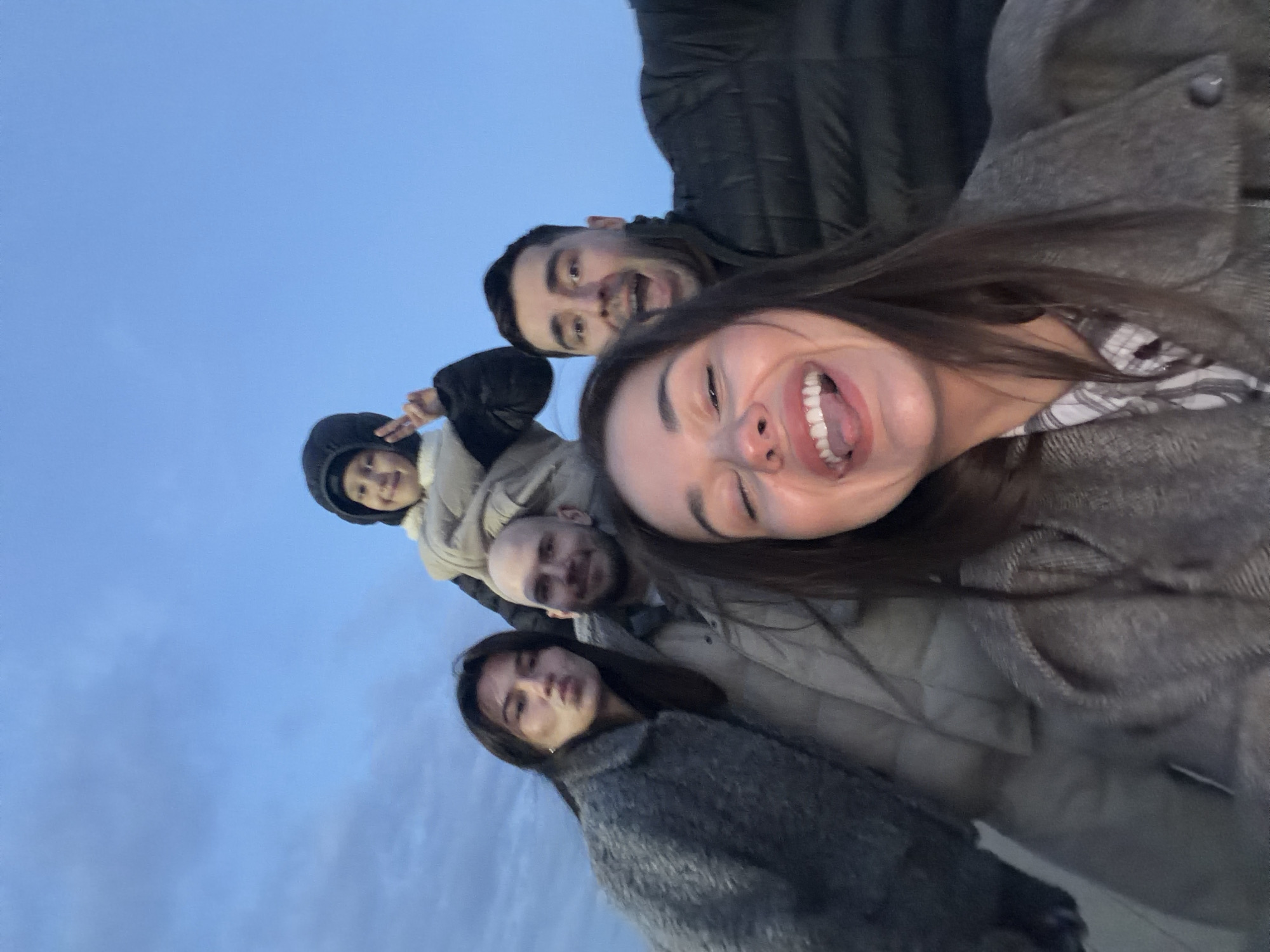 Five individuals are posing for a selfie against a blue sky backdrop. The composition is tilted. The foreground features a woman with long brown hair, wearing a gray coat, smiling widely and opening her mouth. Behind her, a bald man in a light-colored jacket smiles. A person wearing a black hooded jacket is behind him. Further back, another man smiles, clad in a dark jacket. The last person in the row is a woman with long dark hair, wearing a gray coat. The image gives the impression of the group standing somewhere, with the camera pointing upwards and slightly at an angle, with the city of Krasnoyarsk, Russia as the general location of the photograph.
