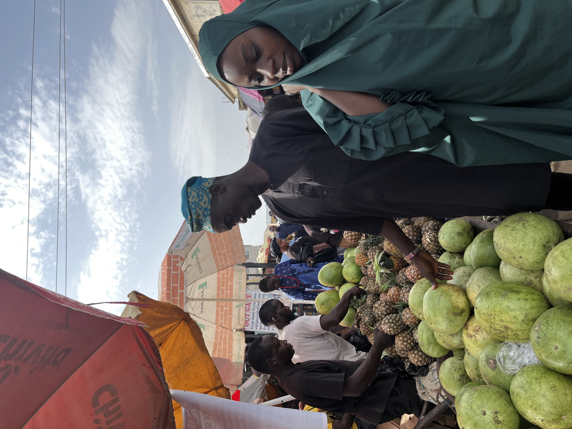 This image depicts a vibrant outdoor market scene in Rafin Par, Nigeria. The foreground is dominated by a large display of green watermelons, with some partially obscured by pineapples in the background. Several people are present, including two men on the left appearing to be vendors or customers inspecting the fruit. One man wears a dark t-shirt, and the other a white t-shirt. A man in the center, wearing a dark traditional outfit and a blue patterned cap, is also looking at the produce. To the right, a young woman in a dark green hijab is partially visible, with a portion of her face showing as she looks down. Behind the main group, other market-goers and stalls can be seen. The scene is set under a bright sky with scattered clouds, with colorful umbrellas providing shade. A sign with text is visible on one of the umbrellas. The overall impression is one of a bustling local market.