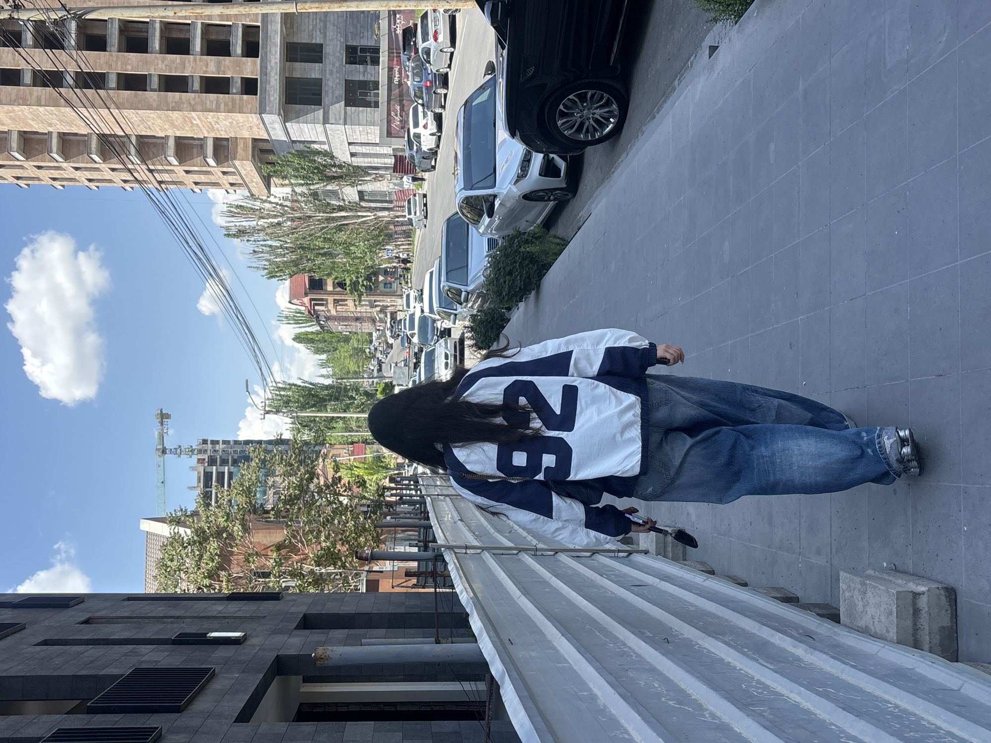 A street scene in Yerevan, Armenia shows a long-haired person wearing a white and navy blue jacket with the number "26" on the back, loose-fitting jeans, and silver shoes, walking along a concrete sidewalk. The person is facing away from the camera and appears to be holding a smartphone. A corrugated metal fence runs along the lower part of the frame. The sidewalk is lined with parked cars on the right. In the background, there are buildings of various heights and architectural styles, including a tall building under construction with a green crane visible. The sky is blue with scattered white clouds. Overhead power lines are visible.