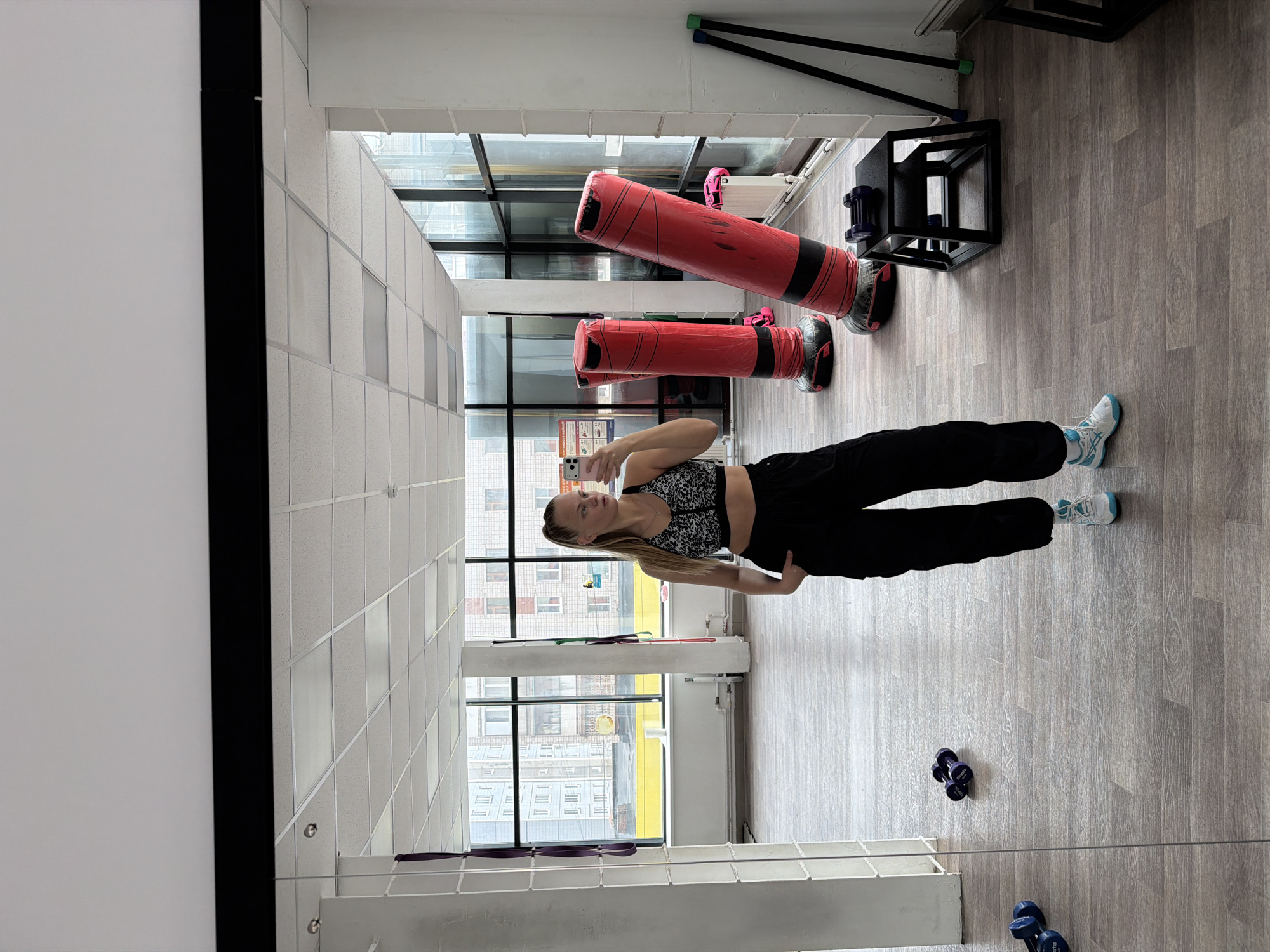 A woman is taking a selfie in a gym with two red punching bags, a black platform, and dumbbells. Outside the windows is a view of buildings in Izhevsk, Russia.