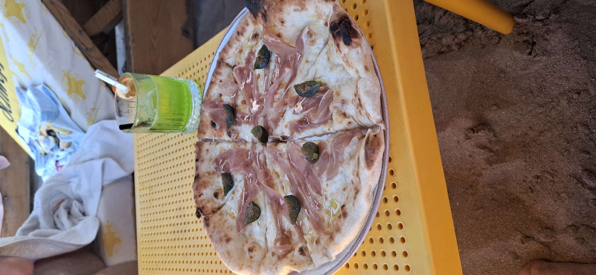 This media file shows a close-up, overhead view of a pizza and a green beverage on a yellow, perforated table. The pizza appears to be a thin-crust variety, topped with slices of prosciutto and green olives or capers, and appears to have been cut into four slices. A glass of bright green liquid, likely a cocktail or juice, sits to the left of the pizza, with a straw and a slice of citrus fruit visible. The table is set on a sandy surface, with some fabric items, possibly towels or beachwear, visible in the background to the left. The sand and fabric suggest an outdoor, possibly beachside, setting. The lighting suggests it is daytime, with no specific time of day discernible. The overall mood is relaxed and informal. There is no visible text in the media file. The location context is Sozopol, Bulgaria, which aligns with the beachside setting.