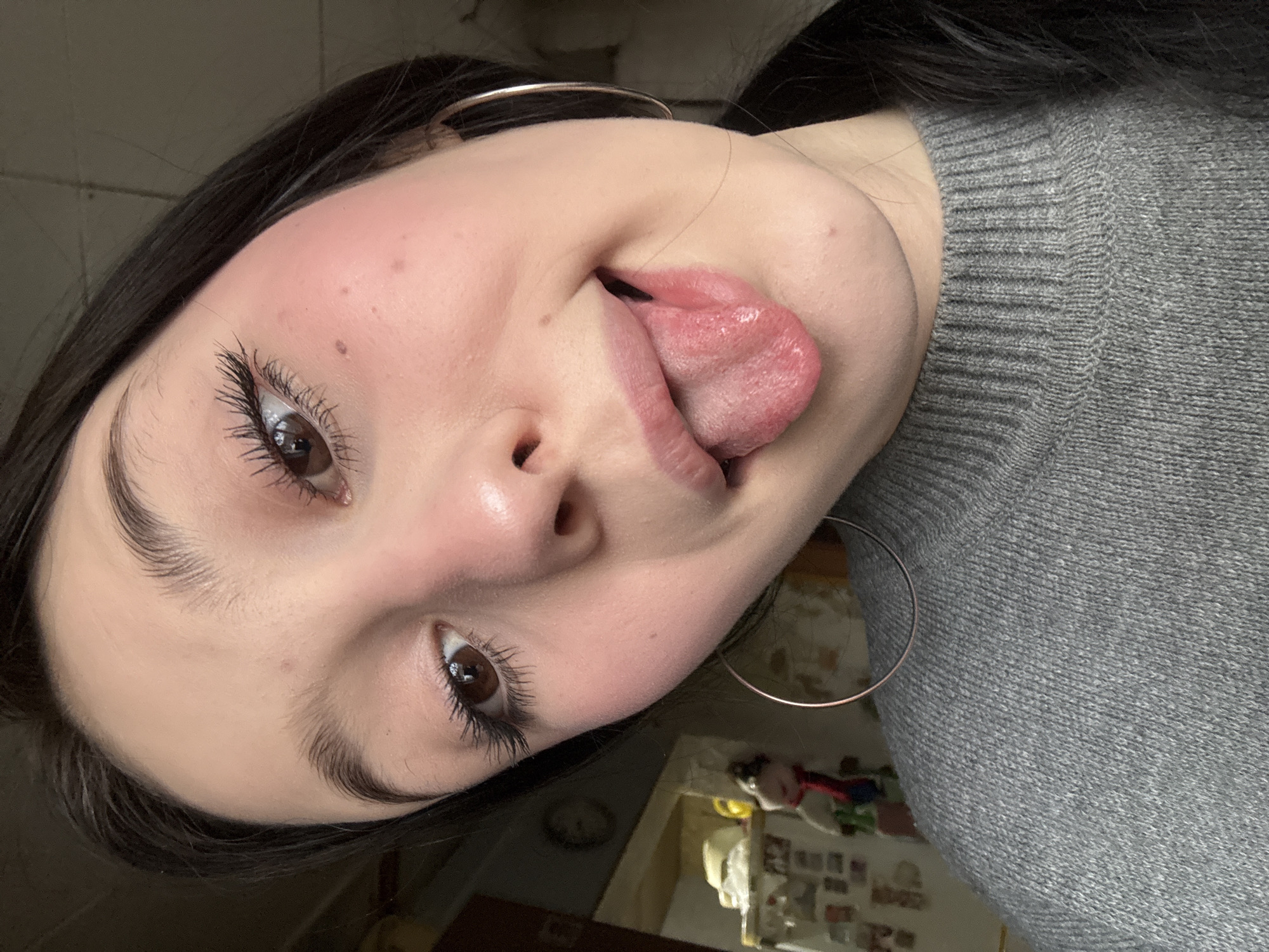 A close-up photograph shows a young woman with dark hair and brown eyes, wearing a gray sweater and large hoop earrings. She is sticking her tongue out. The background appears to be a domestic interior, with glimpses of furniture and other objects. There is no indication of the city of Фоки, Russia.