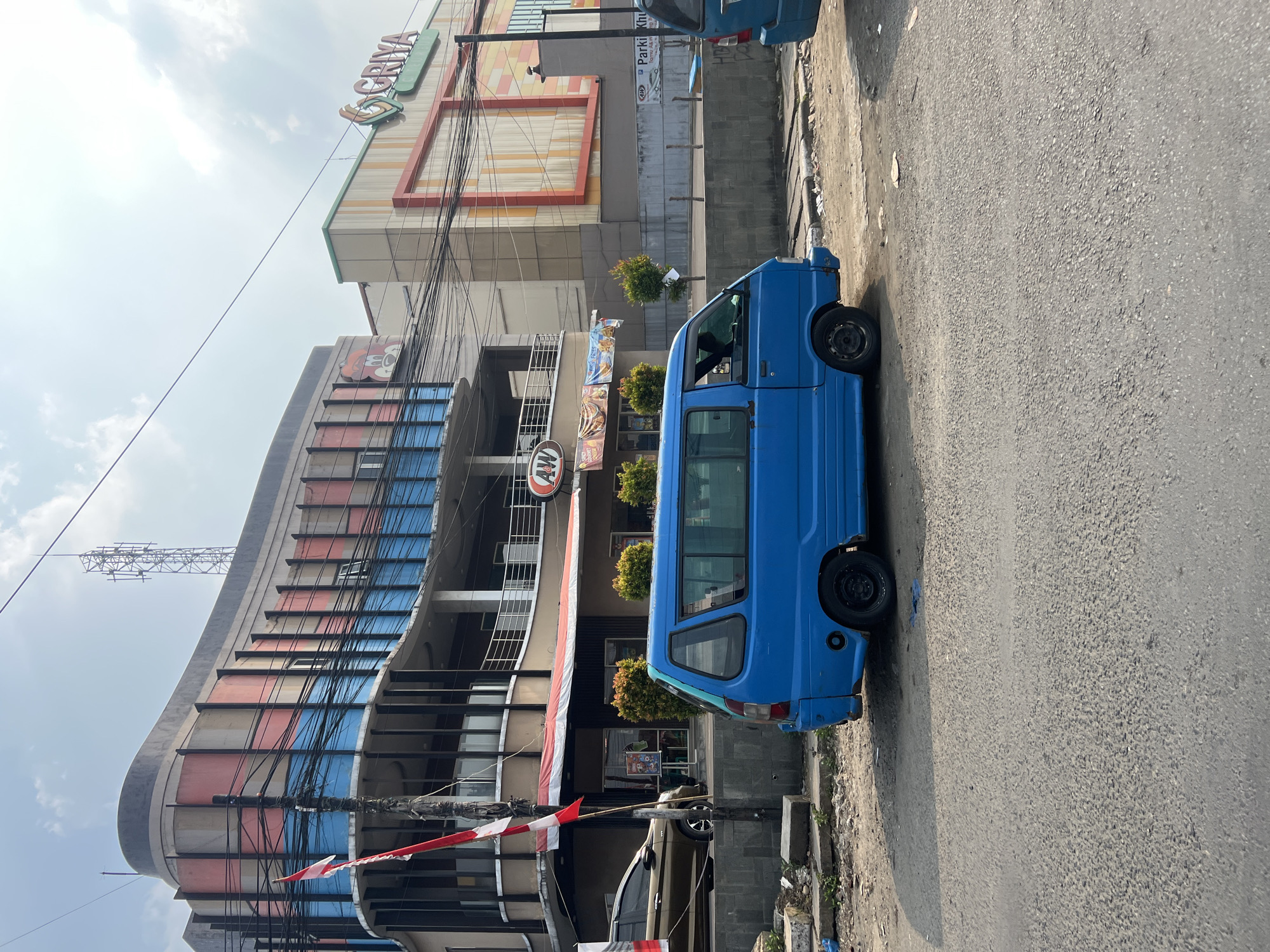 A bright blue minivan is parked on the side of a street in Citeureup, Indonesia. The minivan is positioned to the left of the frame, facing towards the right. In the background, a large building complex is visible. The building on the left has a rounded facade with horizontal bands of pink, grey, and blue. A red and white Indonesian flag is attached to a pole in front of this building. To the right of this building is another structure with a prominent sign that reads "GRIYA" in green and orange letters. The sky above is partly cloudy. The street in the foreground appears to be asphalt, with a dirt and concrete area bordering the building. There is a cell tower visible on top of the left building.