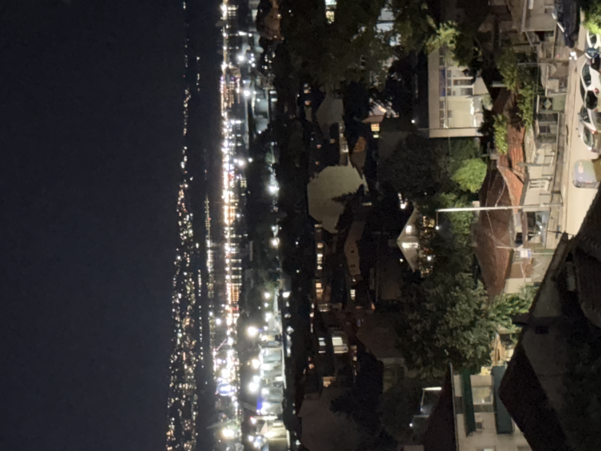 The image shows a nighttime view of a city with buildings and lights. In the foreground, there are several houses with illuminated windows. Some of the houses have tiled roofs and are surrounded by trees. Further in the background, there is a densely lit urban area stretching towards the horizon, with numerous lights reflecting on what appears to be a body of water. The sky is dark and clear, indicating a night scene. The overall impression is of a residential area overlooking a larger, more illuminated part of the city. There are no people or specific actions clearly depicted in the image.