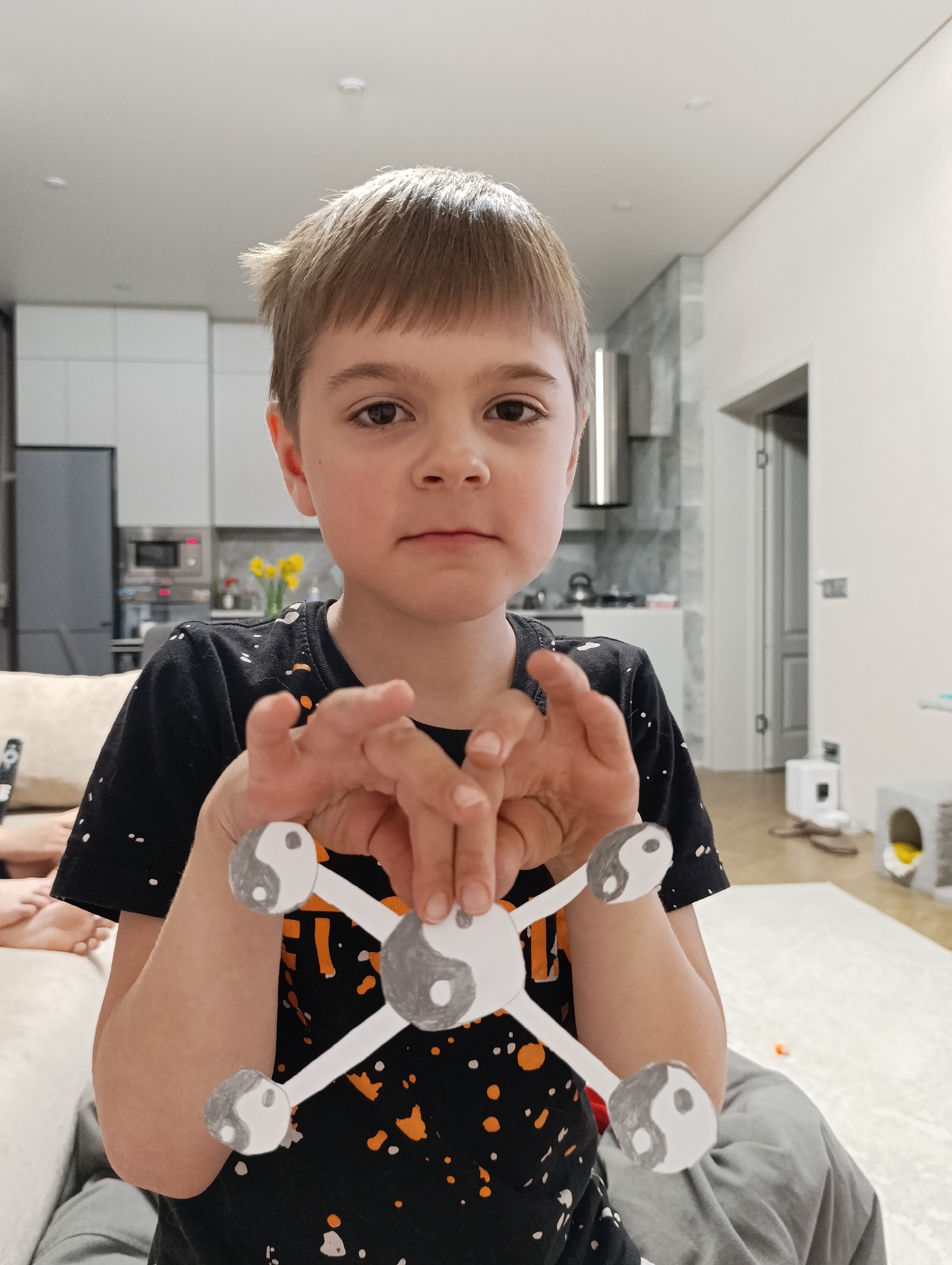 A young boy is holding up a paper craft. The craft consists of a central circle connected to four arms, each ending in a yin-yang symbol. The boy is fair-skinned with brown hair. He is wearing a black t-shirt with white and orange splatters. The setting appears to be indoors, possibly a home. A kitchen is visible in the background, with white cabinets, stainless steel appliances, and yellow flowers on a counter. Part of a couch with a person on it is visible to the left, and there is a cat house or bed visible in the background. There is no information in the image to confirm that it was taken in Chaikovsky, Russia.