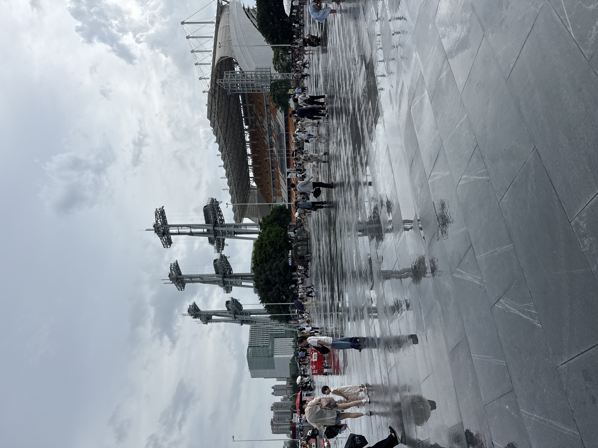 The image shows a wide, wet, paved plaza in front of a stadium, likely in Tianhe District, China, under a cloudy sky. The wet surface of the plaza reflects the surrounding environment and the people walking on it. Several individuals are visible, some holding umbrellas, suggesting it might be raining or has recently rained. In the background, a large stadium with orange seating and a modern architectural design is prominent. Towering over the plaza and stadium are massive metal structures, possibly part of the stadium's infrastructure or nearby industrial equipment, with a complex lattice design. A cluster of trees can be seen to the right of the stadium, and beyond that, urban buildings line the horizon. The overall atmosphere is somewhat subdued due to the overcast weather and wet conditions.
