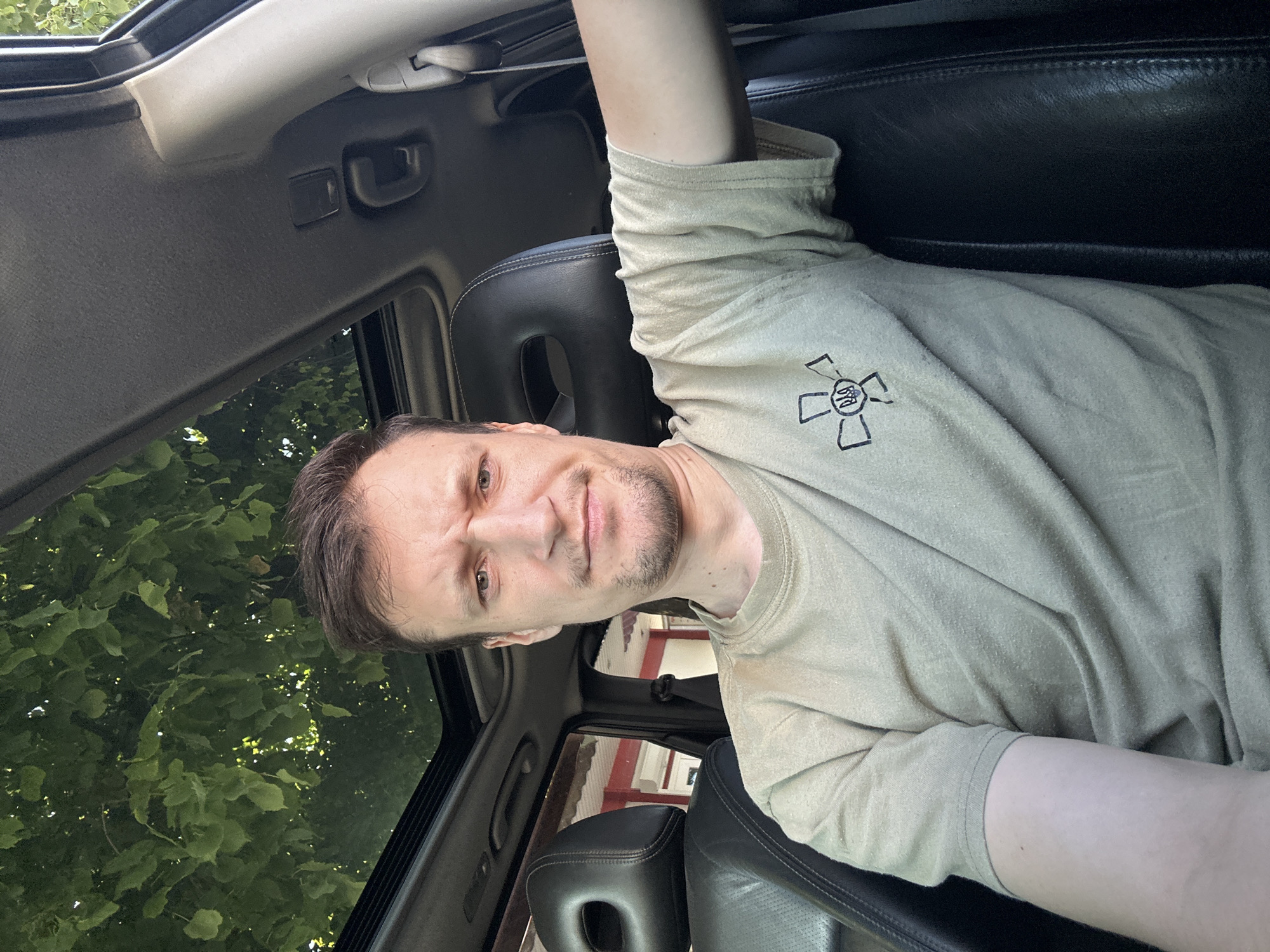 A man with short brown hair and a light beard is sitting in the driver's seat of a car, looking directly at the camera with a concerned expression. He is wearing a light brown t-shirt with a symbol on the left chest. The car has a black leather interior and a sunroof through which green foliage is visible. The background outside the car appears to be buildings. The lighting suggests it is daytime, possibly late afternoon. The location is identified as Kamianets-Podilskyi, Ukraine.