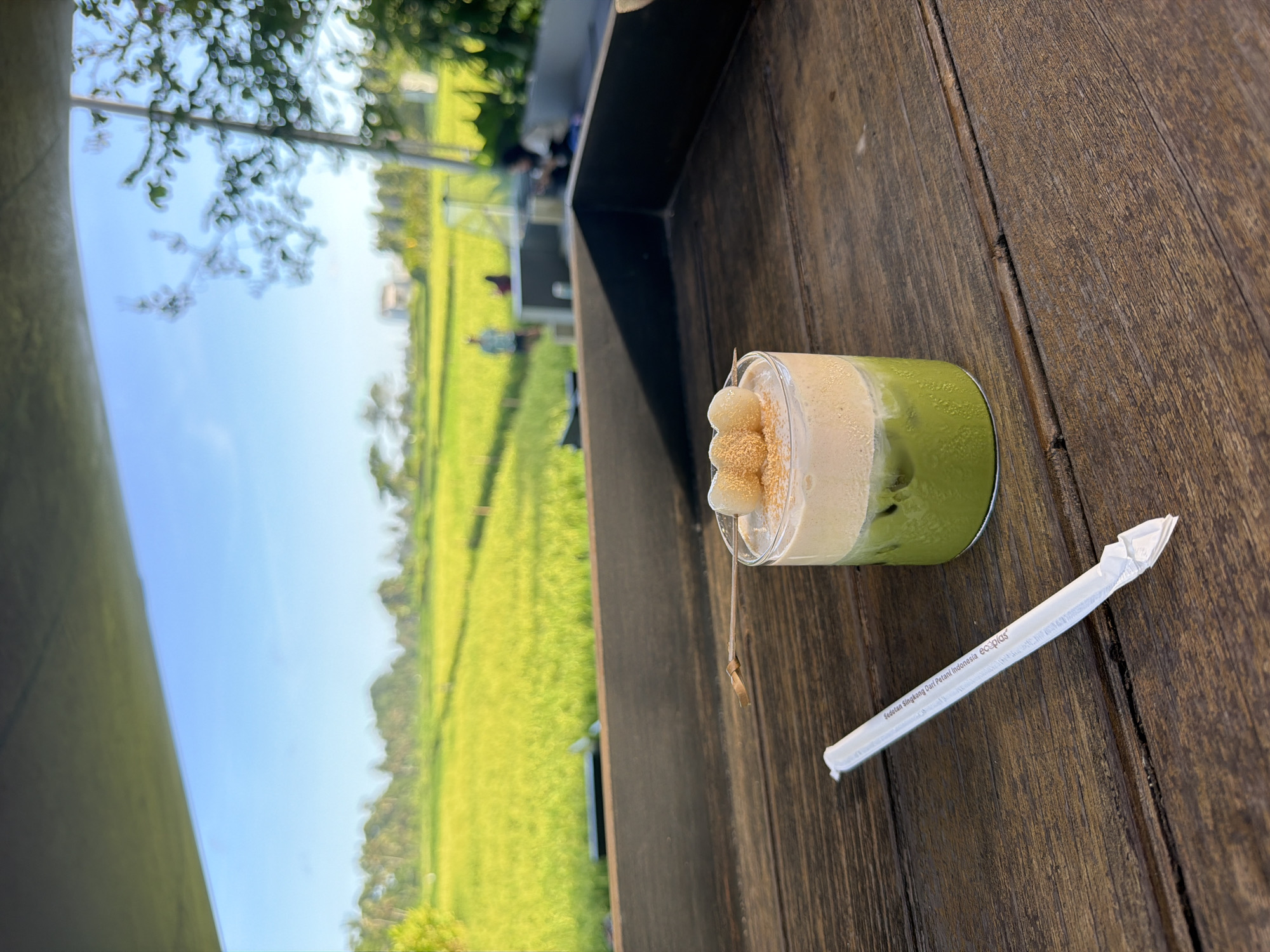 Here is a detailed description of the image:
**Scene:** An eye-level, outdoor shot taken in daylight, seemingly on a wooden table. In the center right of the frame sits a glass containing a green-colored beverage (possibly matcha or iced green tea) topped with a light-colored foamy layer and what appears to be small, tan-colored rice cakes or mochi. A wrapped drinking straw lies to the right of the glass on the wooden surface.
**Environment:** The background, slightly blurred, shows a rural landscape with lush green rice paddies. A few indistinct figures of people can be seen in the distance. The weather appears sunny and clear, indicated by the bright blue sky.
**City:** Based on the rice paddies and likely beverage style, the location is inferred to be in the area of Sukawati, Indonesia.