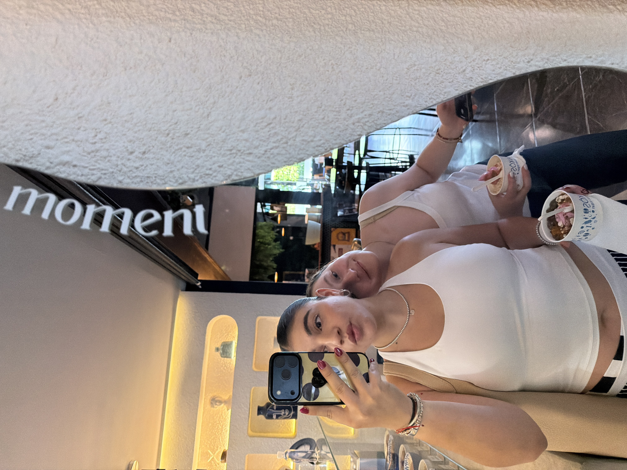This image captures a mirror selfie of two young women relaxing in a modern commercial space, likely a dessert shop or cafe in Puebla, Mexico. The photo is taken from a reflective surface, possibly a large wall mirror or ceiling, looking down at the subjects.
In the foreground, a woman with dark hair, pulled back, holds an iPhone with a black and white polka-dotted case in her right hand to take the picture. She is wearing a white tank top, a delicate silver beaded necklace, and several bracelets, including a red string and silver beaded ones. Her fingernails are painted a dark red, and she has a slight pout.
Behind her, another woman reclines comfortably, looking towards the camera's reflection with a faint smile. She also wears a white top and holds a white cup filled with a dessert, likely yogurt or ice cream, topped with pink, brown, and white additions, with a spoon inserted. A similar dessert cup is partially visible next to the foreground woman, bearing the partial text "2OY."
The reflected background reveals a contemporary interior with sleek, illuminated display shelves showcasing small items, possibly jewelry or decorative objects. Through a reflected window, greenery and parts of other buildings are visible, suggesting it is daytime. The word "moment" is prominently reflected, appearing upside down and backward, indicating signage within the establishment. The scene conveys a relaxed and casual atmosphere as the friends enjoy their treats and capture the "moment."