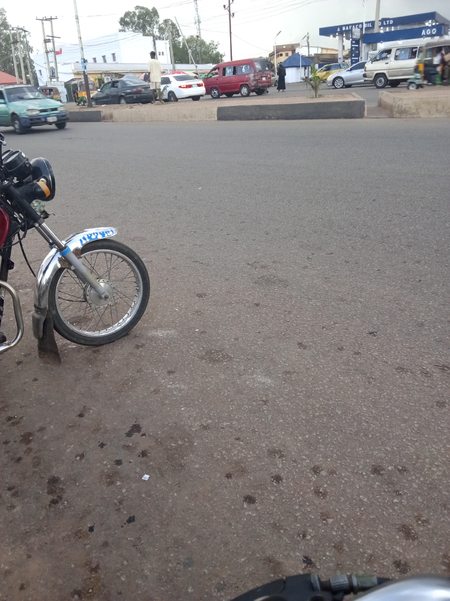 This is a street-level photograph taken in Jos, Nigeria, likely during daylight hours with overcast skies. The foreground features the front wheel and part of the frame of a motorcycle. The motorcycle appears to be a standard street bike, with a chrome fender and spokes visible on the wheel. The background depicts a busy street scene with several vehicles.
On the left, a light green saloon car is driving past. Further down the street, a dark sedan is parked. A white saloon car and a red minivan are also present. In the distance, a white van, possibly a bus, and a yellow saloon car are visible. A white structure with a blue awning and the text "BASACO OIL LTD" is clearly visible, indicating a fuel station. People are seen near the vehicles, including a man in a light-colored outfit standing on the roadside and individuals near the fuel station. The environment includes a concrete median separating lanes of traffic and a paved road surface with some scattered dark spots, possibly oil stains. Utility poles with electrical wires are also present.