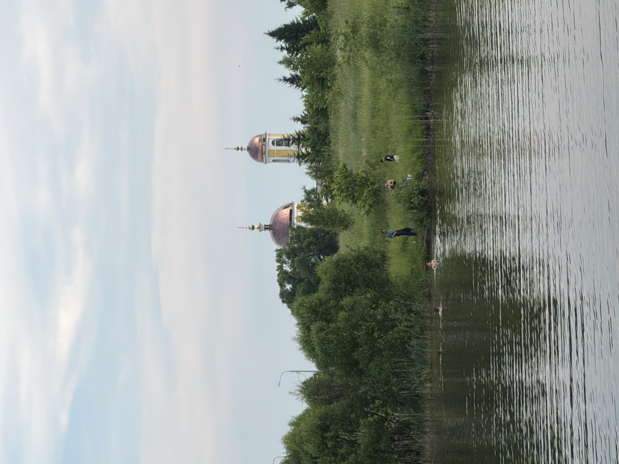 In Satino-Russkoe, Russia, a serene lake scene unfolds. The foreground is dominated by the calm, rippling water of the lake. In the mid-ground, a lush green bank is dotted with trees, reeds, and tall grass. Several people are visible on the bank and in the water. On the right side of the bank, a man stands dressed in a blue shirt and dark pants. Further to the left, a group of three individuals, two women and one man, are gathered, with one woman wearing a dark top and light shorts. In the water, three people are swimming, with one partially submerged near the right bank. In the background, two distinct church domes, one copper-colored and the other golden-capped, rise above the treeline, indicating a religious structure. The sky is overcast with soft, diffused clouds.