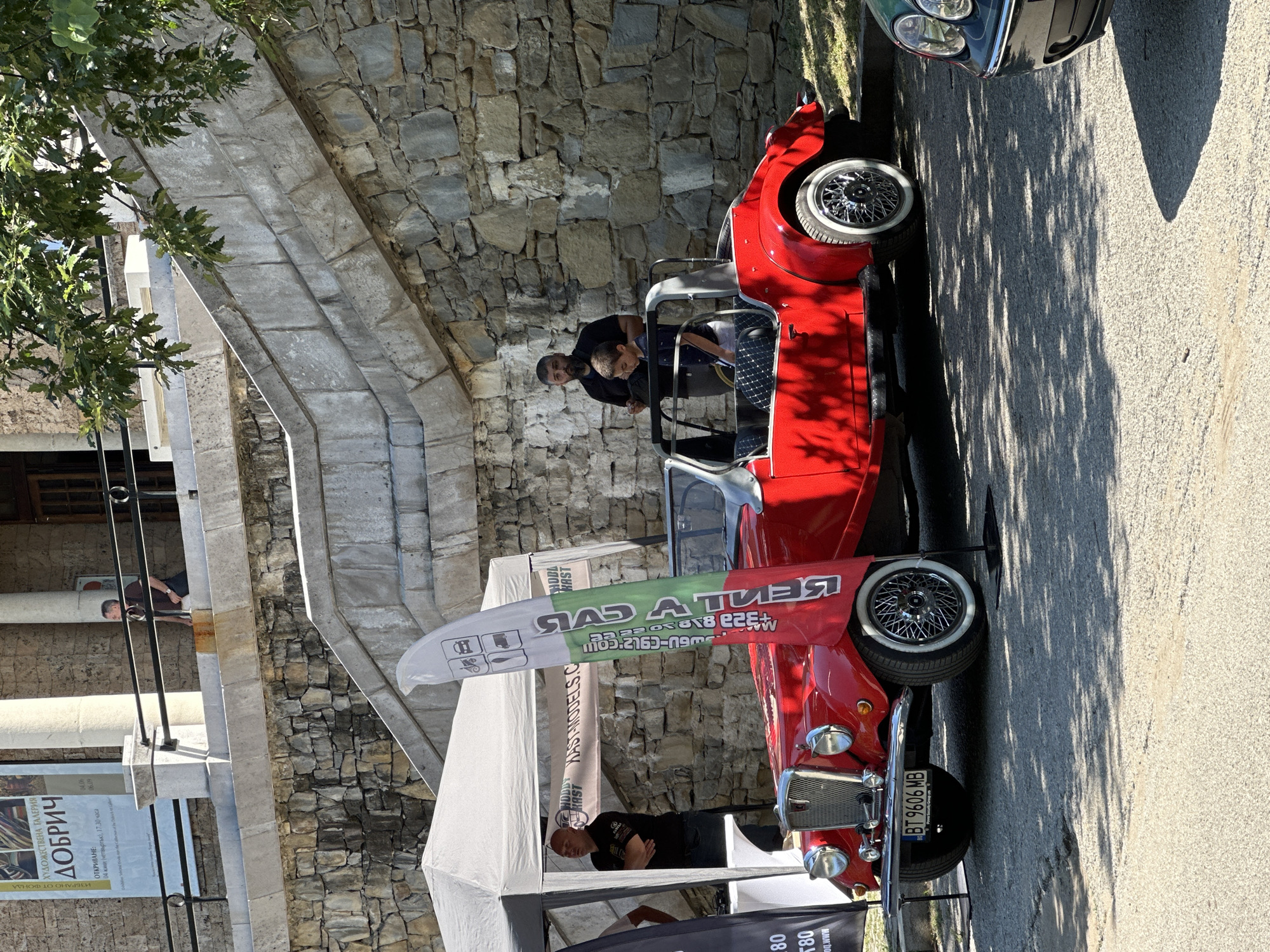 The image captures an outdoor scene in Veliko Tarnovo, Bulgaria, with a vintage red convertible car as the central subject. The car is parked on an asphalt surface dappled with sunlight and shadow from overhead trees. To the left, a white tent and a black flag are visible. A prominent flag with Bulgarian colors and various icons stands behind the car. In the background, a stone wall forms part of a building or structure, with a man in a dark shirt and a young boy standing near the car. Another man is partially visible under the tent on the left. The license plate of the car reads "BT 9606 MB". Above the stone wall, a man is seen leaning over a railing. The overall impression is of an event or exhibition, likely related to vintage cars.