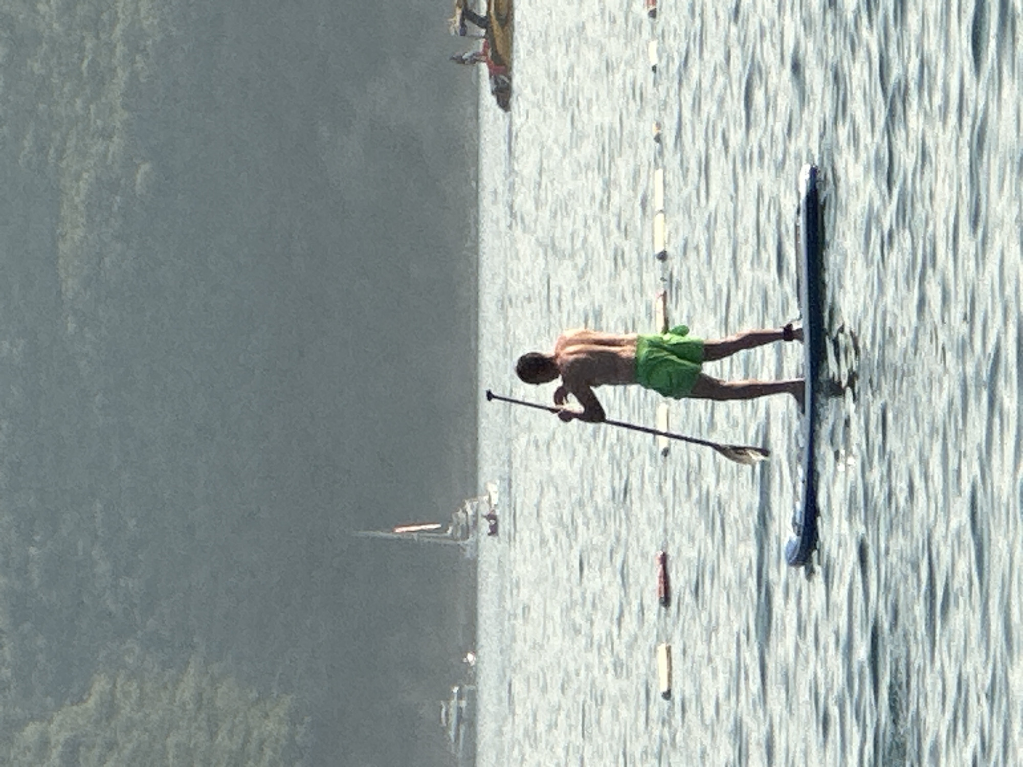 A person is paddleboarding on a body of water, holding a paddle. The person is wearing green shorts. In the background, there are several boats, including a sailboat, and a forested hill. There are also several buoys in the water. The scene appears to be a recreational area. The location is the city of Boreti, Montenegro.