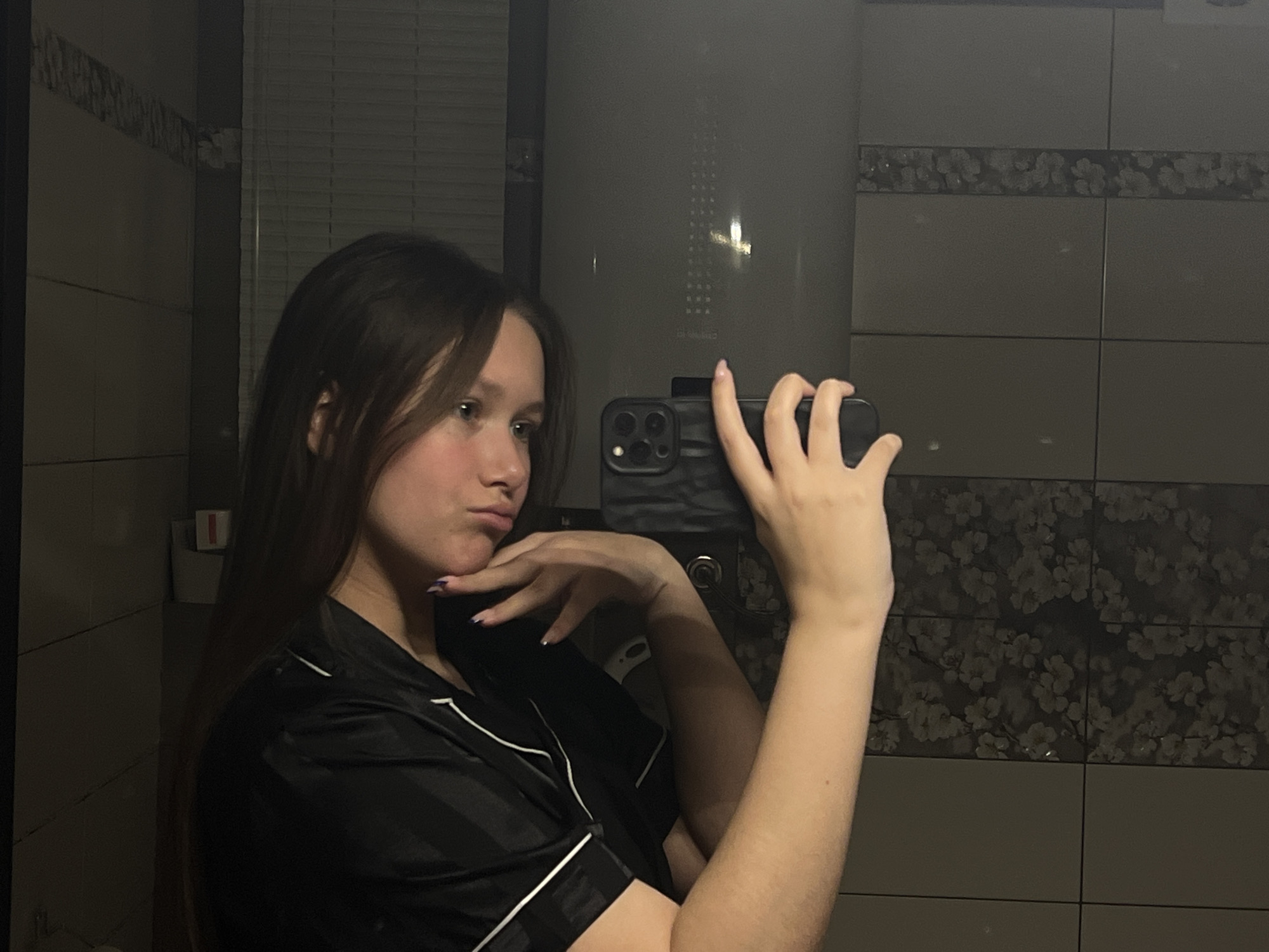 A young woman with long, dark hair is captured in a mirror selfie, taken indoors under dim artificial lighting. She is wearing a black satin pajama-style top with white piping. Her right hand, holding a dark-cased smartphone (likely an iPhone) to take the picture, is visible in the reflection, while her left hand is posed near her chin, contributing to her slightly pouting or thoughtful expression.
The background features dark tiled walls, with a decorative strip of tiles showcasing a subtle floral pattern. Prominently behind the woman is a large, grey cylindrical object, appearing to be a water heater or boiler. Faint text is visible on this appliance, identifying brands such as "ELECTROLUX" and "THERMEX," along with "CHAIKA" (ЧАЙКА in Cyrillic, meaning 'seagull'). Blinds are partially visible in the upper left background, suggesting a window or another reflective surface. The setting, characterized by the tiles and water heater, implies a bathroom or utility room. The dim light suggests it is evening or night. The visible Cyrillic text and type of appliance are consistent with the provided location context of Chaikovsky, Russia.