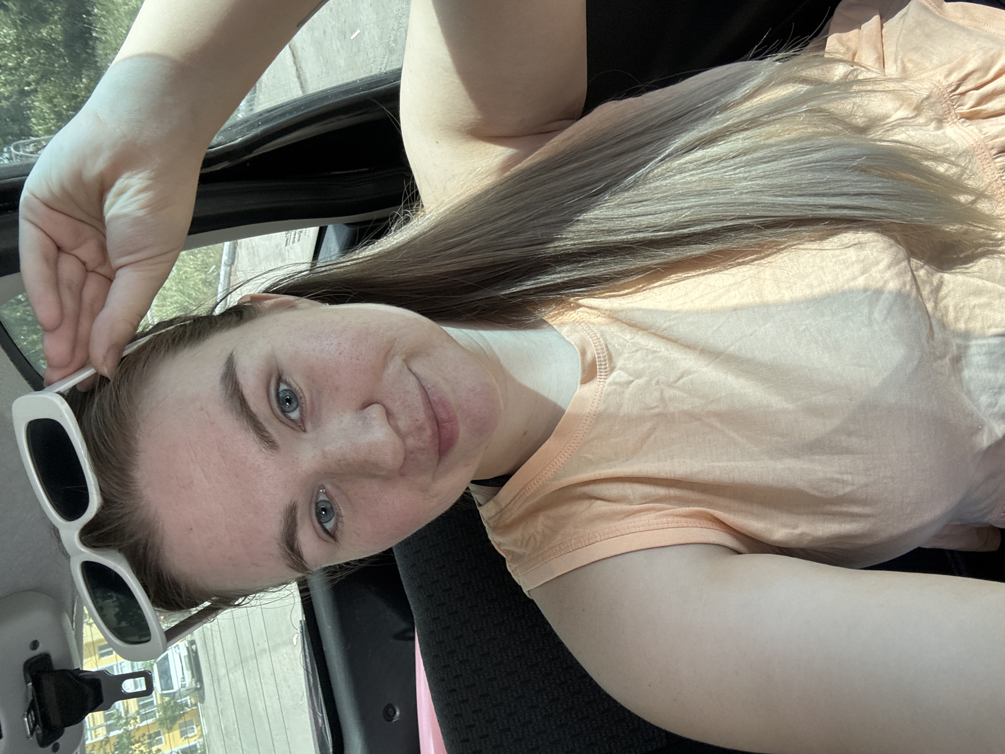 A pale-skinned woman with blonde hair and blue eyes is in a car. She is wearing an orange shirt and holding sunglasses. The location appears to be Chaikovsky, Russia as deduced from objects visible through the car window.