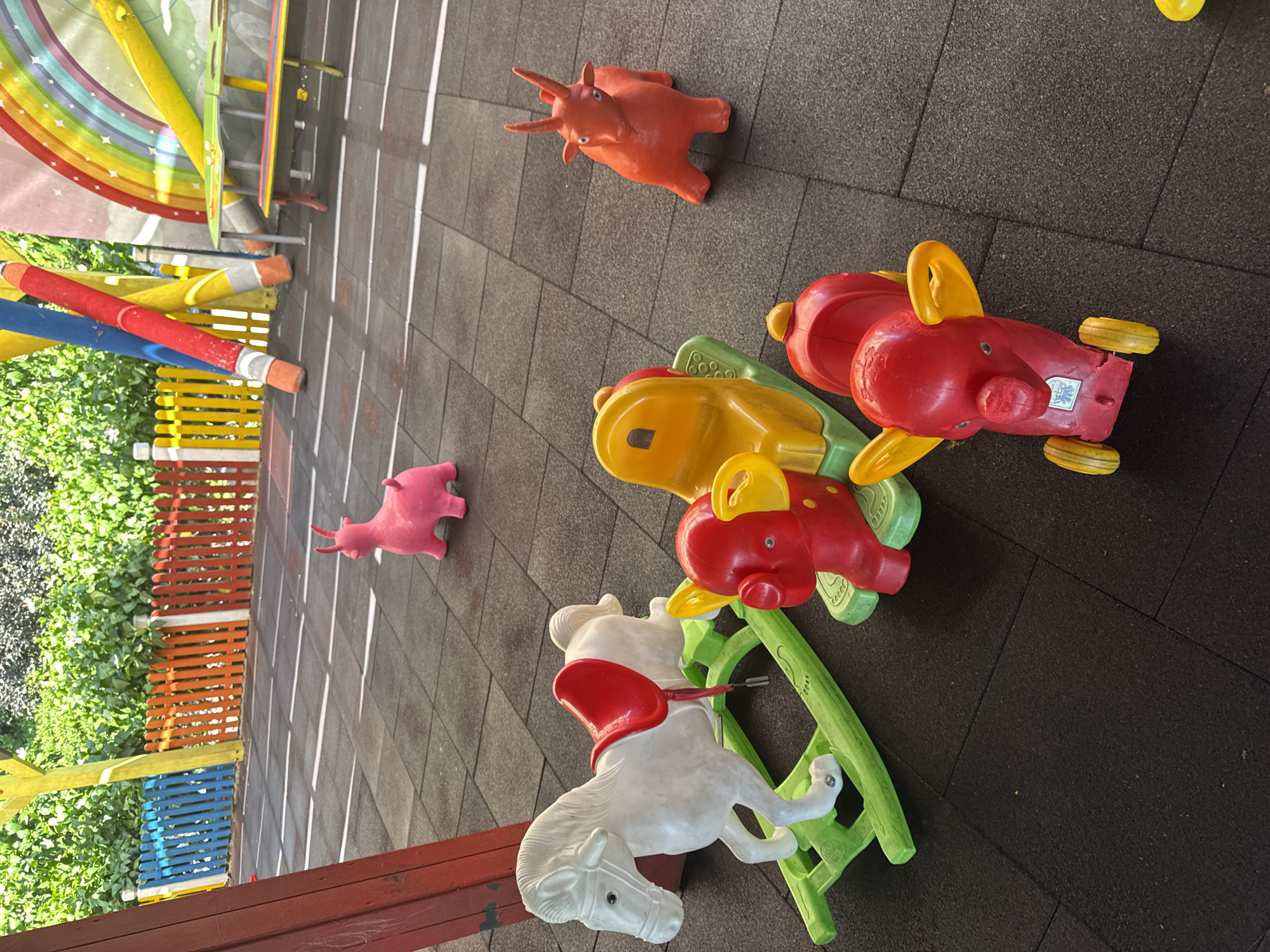 The image depicts an outdoor playground area with a dark grey rubberized ground surface. The playground features several colorful plastic toys, including a white rocking horse with a red saddle, a yellow and green ride-on toy with a pig's face, and another red ride-on toy resembling an elephant. In the background, there is a brightly colored fence, a wooden post, and a section of a rainbow mural. Lush green foliage is visible behind the fence. The lighting suggests it is daytime, with sunlight casting shadows on the ground. The image appears to be taken in Aksu, Turkey, a city known for its tourist attractions and parks. There are no people visible in the image.