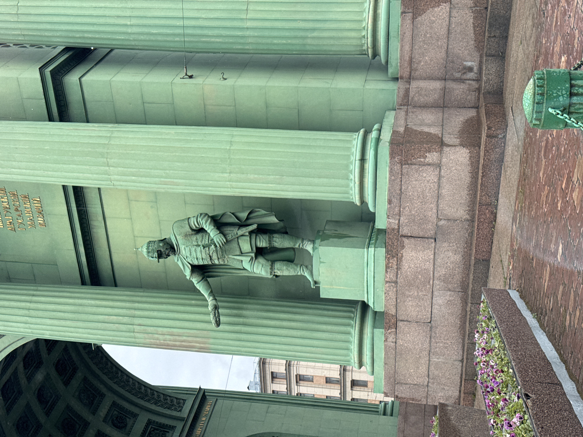 The image depicts a statue of a man in armor standing between green columns of a building with the Alexander Column in the city of Saint Petersburg, Russia. The statue is made of bronze and is wearing a helmet, breastplate, and boots. He has one arm extended. The columns are made of green stone and are fluted. The building has ornate carvings and inscriptions. There is also a planter with flowers near the walkway made of brown paving stones.