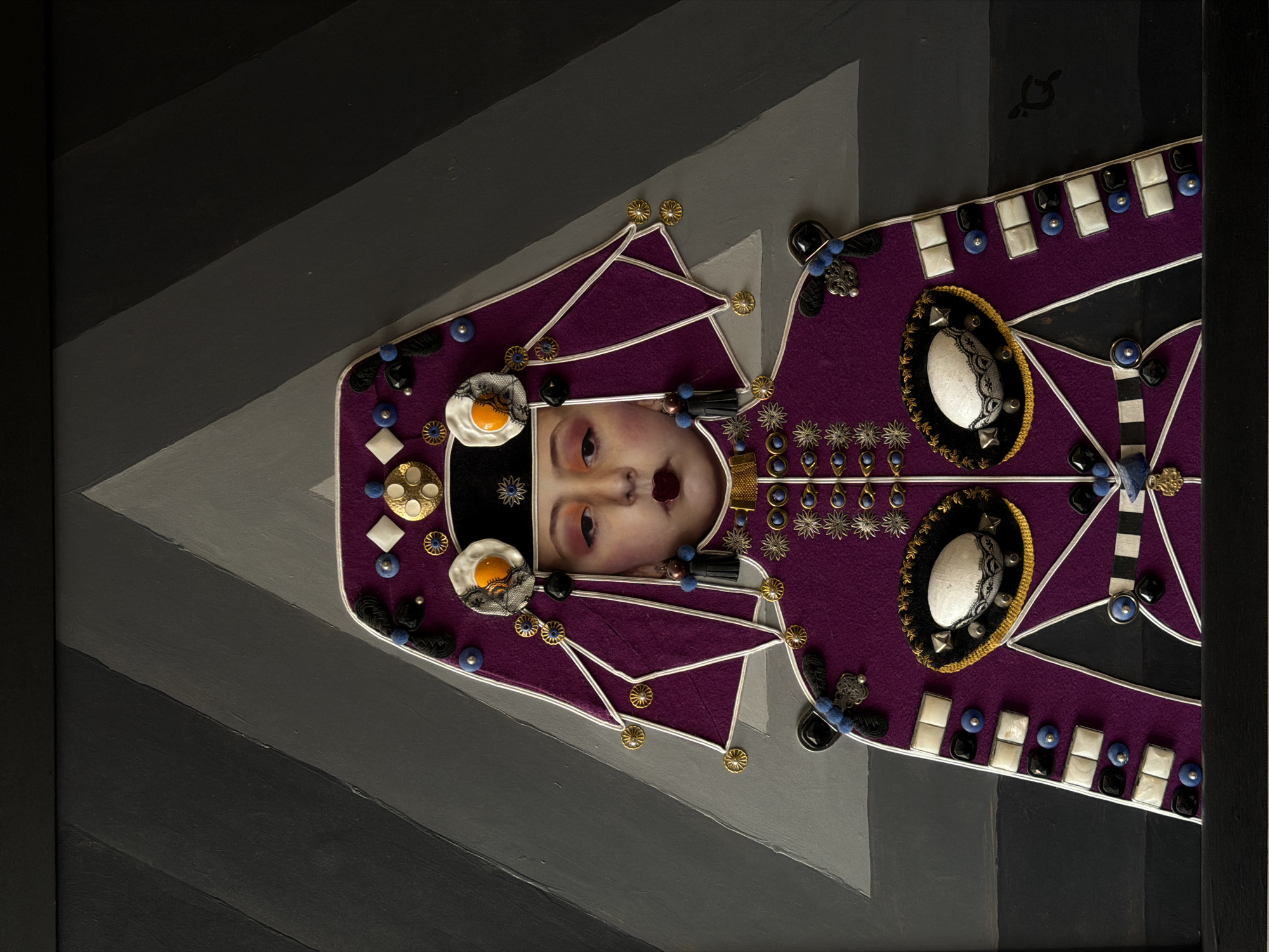 The image is a close-up shot of an art piece. The piece features a central human face with prominent eyes and mouth. The face is surrounded by purple material shaped like a garment, adorned with various embellishments including gold decorations, beads, and what appear to be stylized fried eggs. White cording is used to create a geometric pattern on the purple material. The art is framed and set against a background painted with grey and black angular stripes. The piece gives off a surreal and decorative feel. The information about Yerevan, Armenia is not depicted in the image.