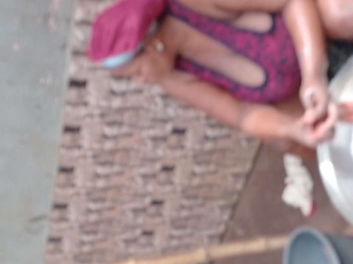 The image displays a blurry, top-down view of a scene, likely indoors or in a covered outdoor area, featuring at least one person. The overall quality is low, making specific details hard to discern.
The central figure appears to be a person, possibly a child, positioned on a patterned surface. They are wearing what looks like a reddish-purple garment, possibly a swimsuit or top, with black or dark-colored details forming a pattern. A bright red or pink cap or hat covers their head. Their body is bent, and their hands are visible on the right side of the frame, seemingly engaged with a light-colored, possibly metallic, bowl or basin. One hand appears to be inside or near the bowl, suggesting an activity involving water or liquid.
The setting consists of at least two distinct floor surfaces. On the left side of the image, there is a smooth, light grey or beige surface, possibly concrete or tiled floor. Adjoining this, and extending under the person, is a textured, patterned surface that resembles brown or beige brickwork, cobblestones, or a patterned mat.
To the right of the person and the bowl, a small white object, possibly a cloth or towel, is visible on the brown surface. In the bottom right corner, a dark grey or blue cylindrical object, resembling a bucket or another container, is partially visible, along with a light brown, linear object that could be a piece of wood or bamboo.
The lighting suggests it is daytime, but no specific time or weather conditions can be inferred from the blurry image. No text is visible.