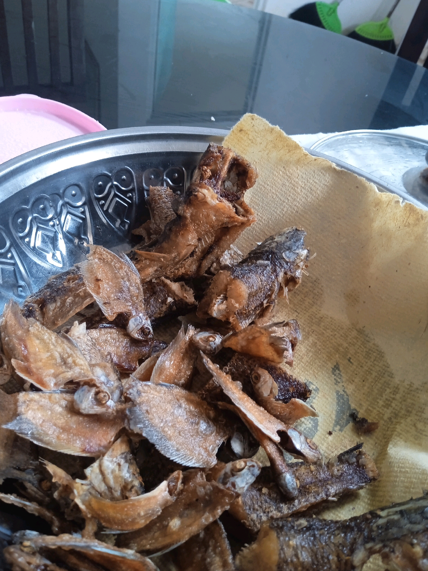 The image shows a close-up of a bowl filled with fried fish. The fish appear to be small, with some whole and others broken into pieces. They are golden brown and crispy, suggesting they have been deep-fried. A piece of brown paper towel is placed inside the bowl, beneath some of the fish, likely to absorb excess oil. The bowl itself is metallic and has a decorative pattern around the rim. In the background, a portion of a glass table and some indistinct objects are visible. The lighting is moderate, highlighting the texture and color of the fried fish. There are no people or active interactions depicted in the image. The location is identified as Banjarbaru, Indonesia.