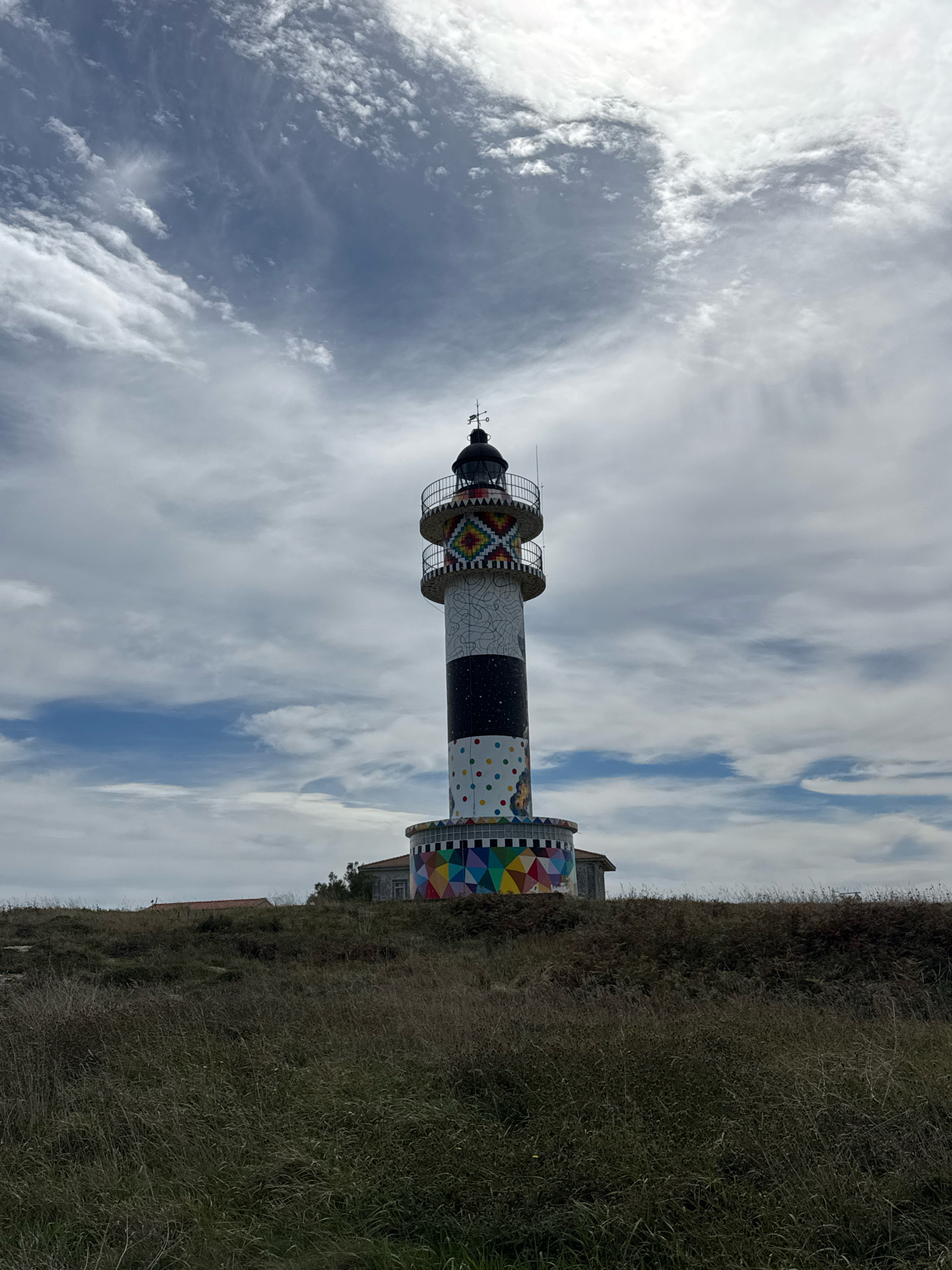 The image shows a tall, cylindrical lighthouse with a black and white striped pattern in the middle section. The top and bottom sections of the lighthouse are adorned with colorful geometric mosaic designs. The lighthouse is situated on a grassy hill, with a cloudy sky overhead. The lighthouse is located in the municipality of Bareyo, Cantabria, Spain, which is known for its coastal landscapes and picturesque lighthouses.
