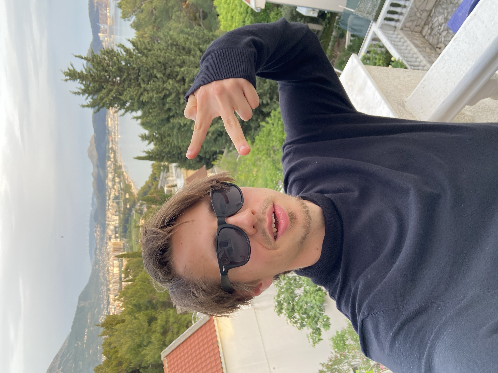 A person, likely male, is posing for a selfie with a background showing part of the city and coast of Šušanj, Montenegro. The person is wearing sunglasses and a dark long-sleeved shirt. Their hand is raised, making a hand gesture with the index, middle, and ring fingers extended. The background includes buildings, trees, and a cityscape. The sky appears overcast. A balcony railing and part of a building are also visible.