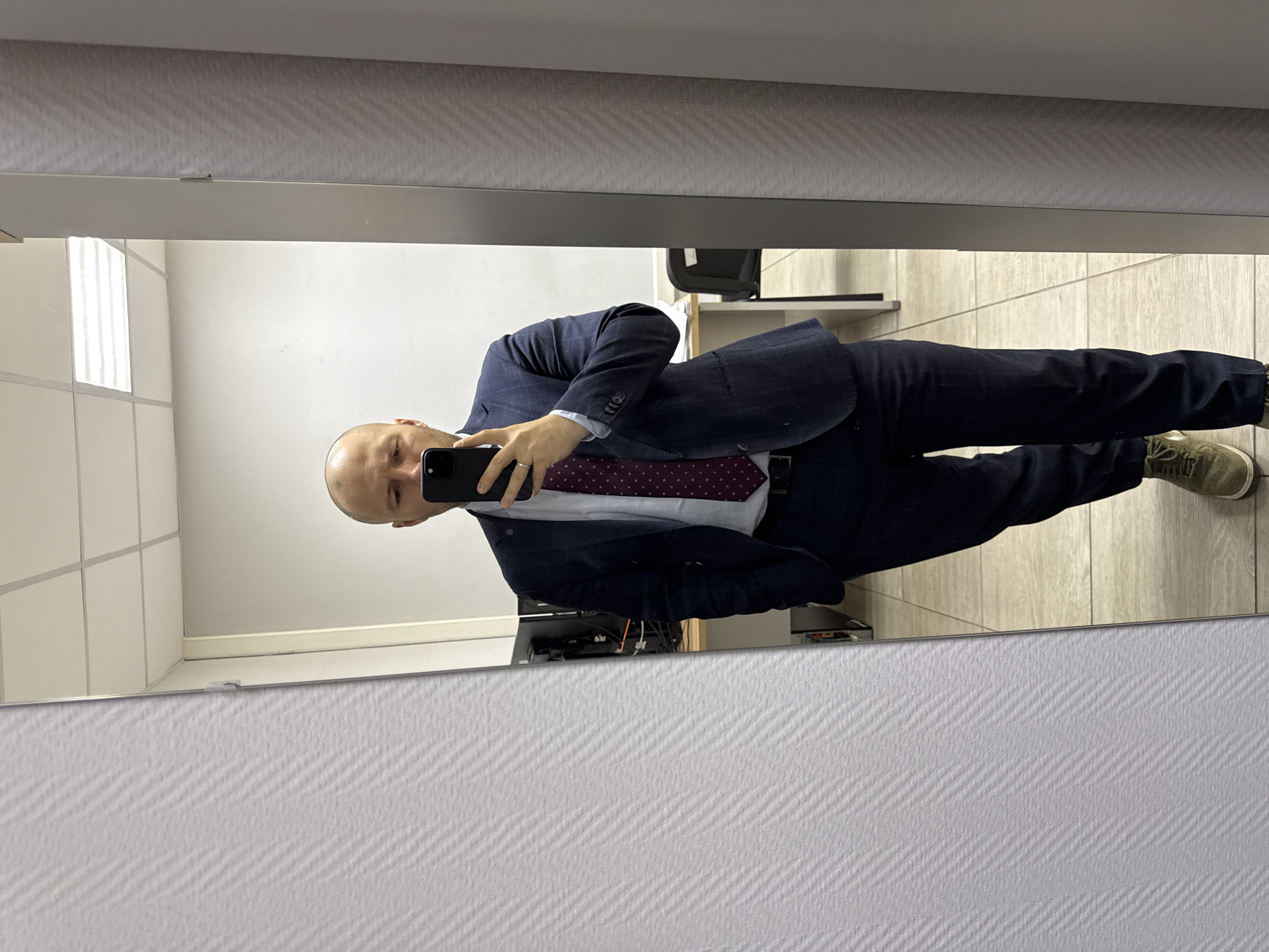 A bald, fair-skinned man in a dark blue suit, white shirt, and patterned red tie is taking a mirror selfie in a room that appears to be an office or workspace. He is holding a smartphone in his right hand, capturing the reflection of himself. The background includes a tiled wall, a shelf with electronic equipment, and a chair. The location cannot be determined from the image alone.