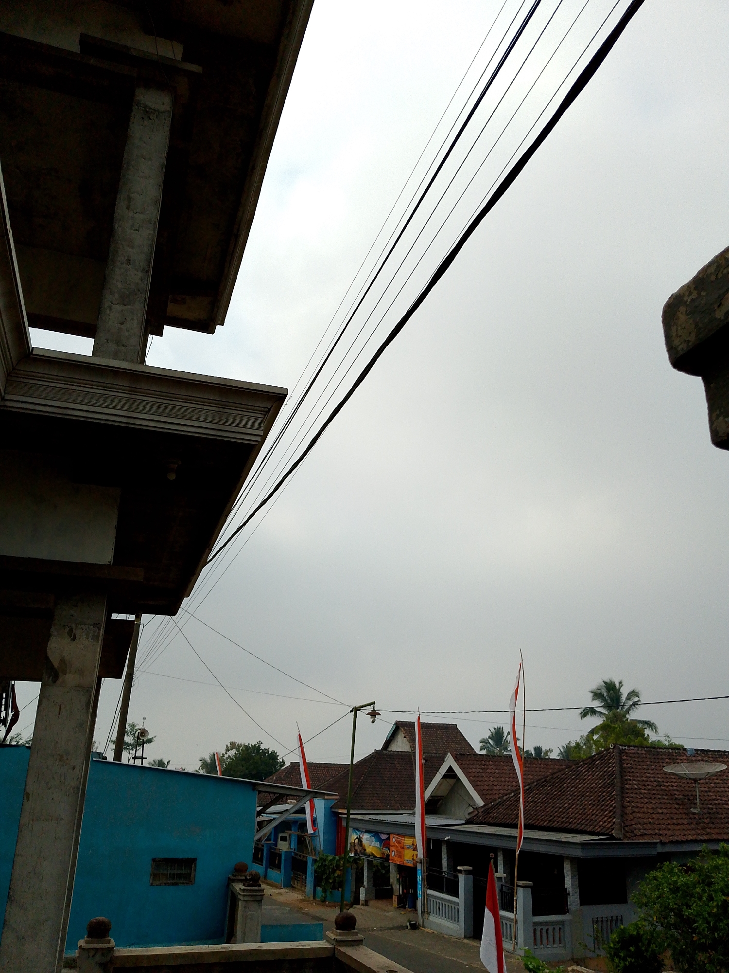 A low-angle outdoor shot captures a street scene in Babadan, Indonesia, under an overcast sky. The foreground is dominated by the dark, unfinished concrete structure of a building on the left, featuring a thick pillar and a decorative cornice. Above, power lines crisscross diagonally from left to right.

The middle ground reveals a street lined with houses. To the left, a vibrant blue building with a small, barred window is visible. Further down the street, houses with terracotta tiled roofs are present. Several Indonesian flags, red and white, are displayed on poles in front of these houses, suggesting a national celebration. A speaker system is mounted on a pole behind the blue building.

The background shows more houses with tiled roofs, interspersed with green trees and a palm tree. The overall atmosphere is quiet and somewhat subdued due to the cloudy weather. No people are directly visible in the image.